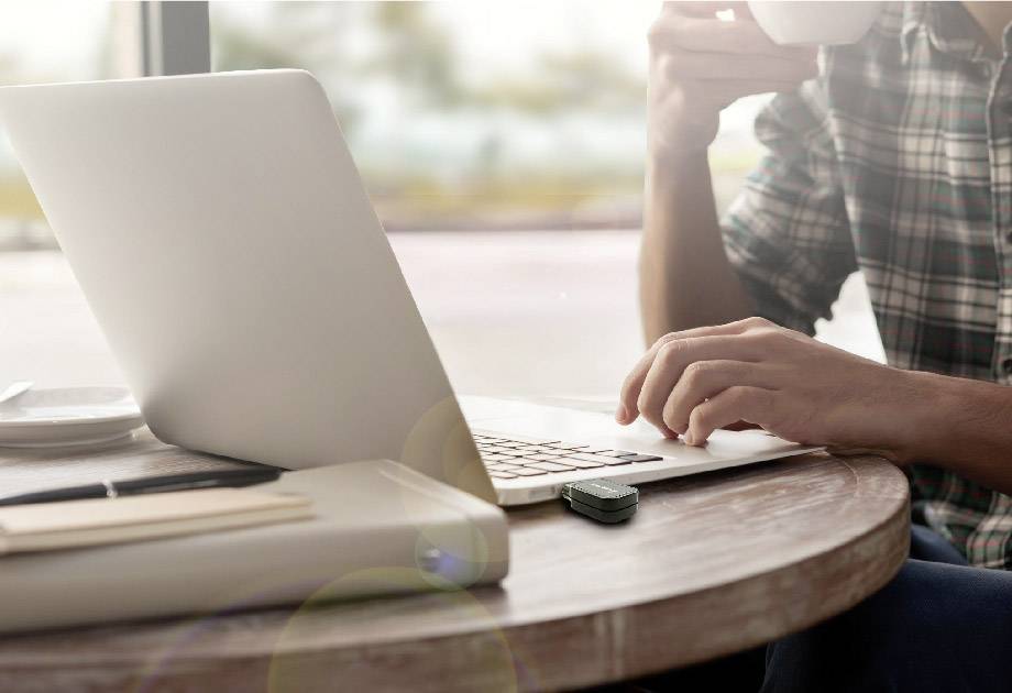 Eine Person tippt auf einem Laptop an einem runden Holztisch und hält eine Tasse. Im Hintergrund ist unscharfe Natur zu sehen.