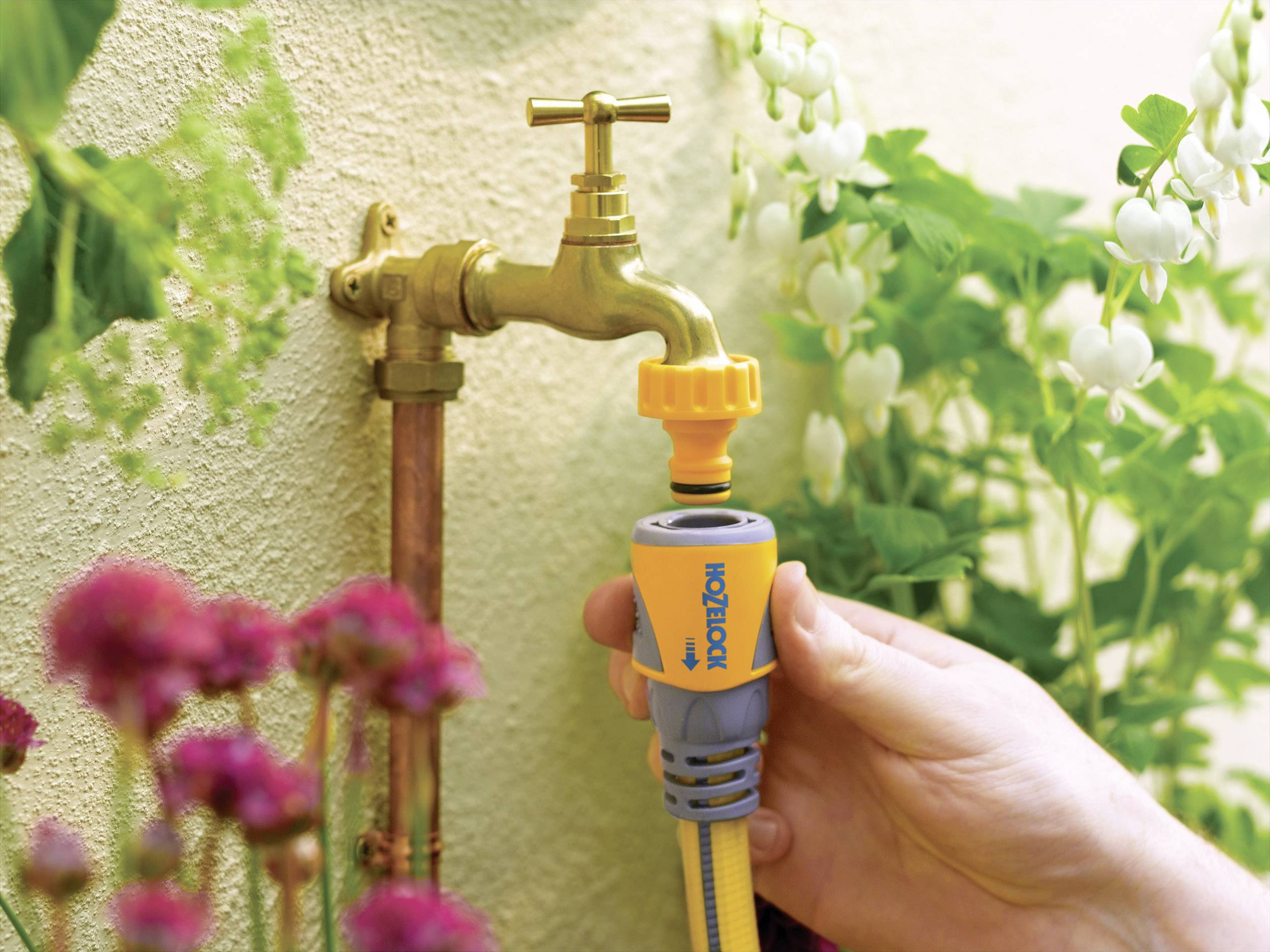 Eine Hand schließt einen gelben Gartenschlauch an einen Außenwasserhahn an. Im Hintergrund sind blühende Pflanzen zu sehen.