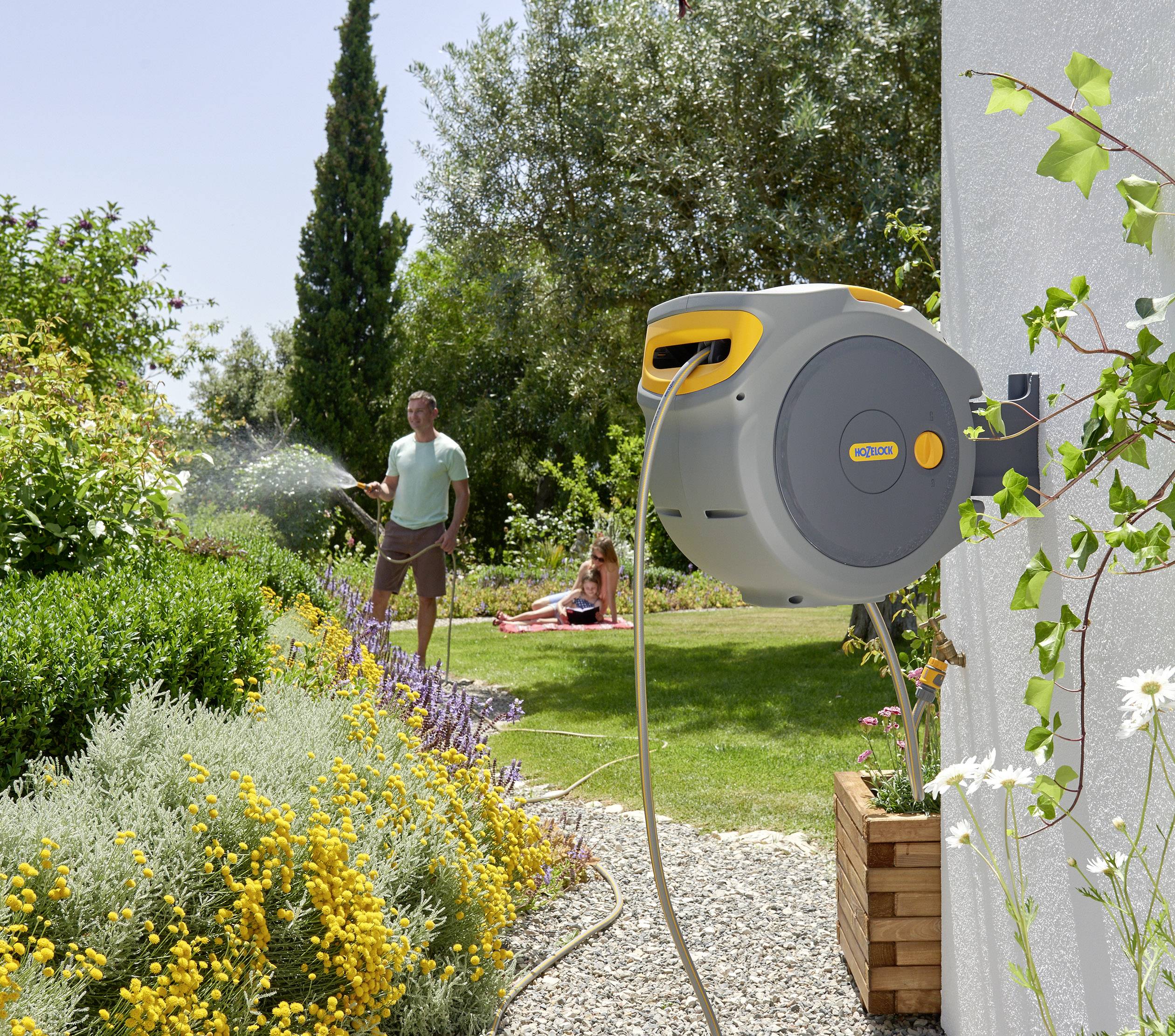 'Ein Mann gießt einen Garten mit bunten Blumen, während ein Kind auf dem Rasen sitzt. Eine gelbe Schlauchtrommel ist an der Wand befestigt.'