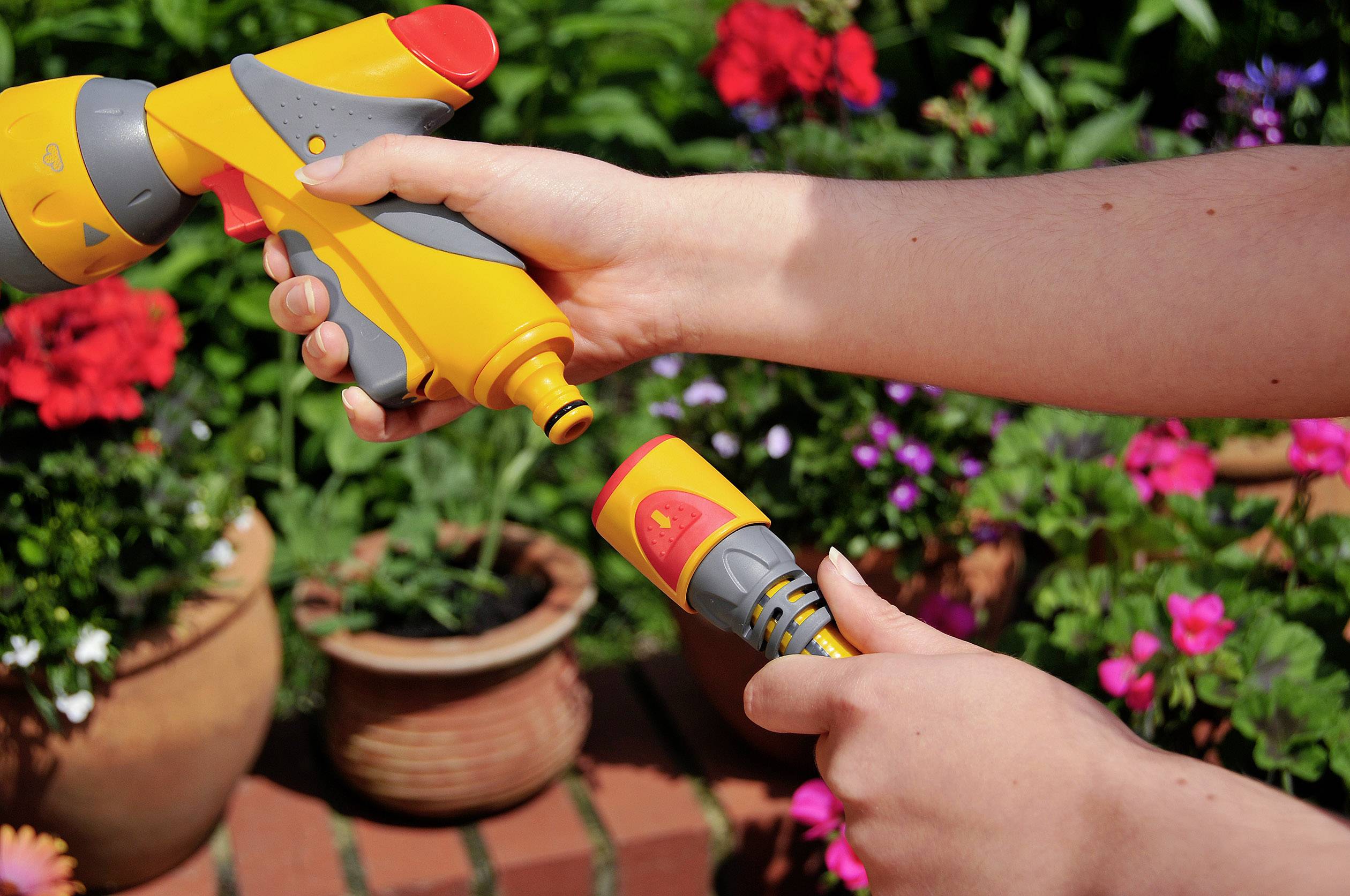 Eine Person hält eine gelbe Gartenschlauchdüse und verbindet sie mit einem gelben Schlauch. Im Hintergrund sind bunte Blumen zu sehen.