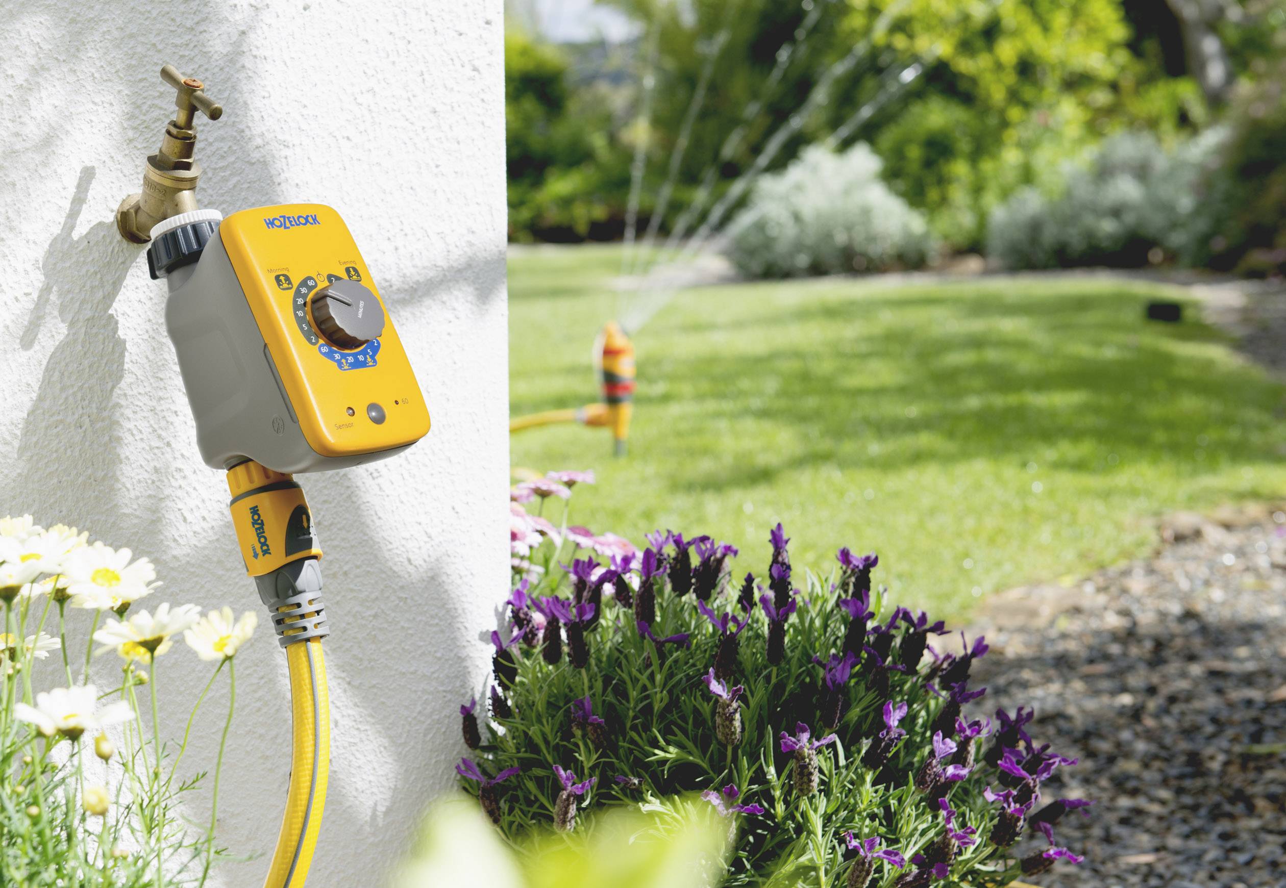 'Gartenbewässerungssystem' zeigt einen automatischen Timer an einem Wasserhahn. Im Hintergrund sprengen Sprinkler einen grünen Rasen.