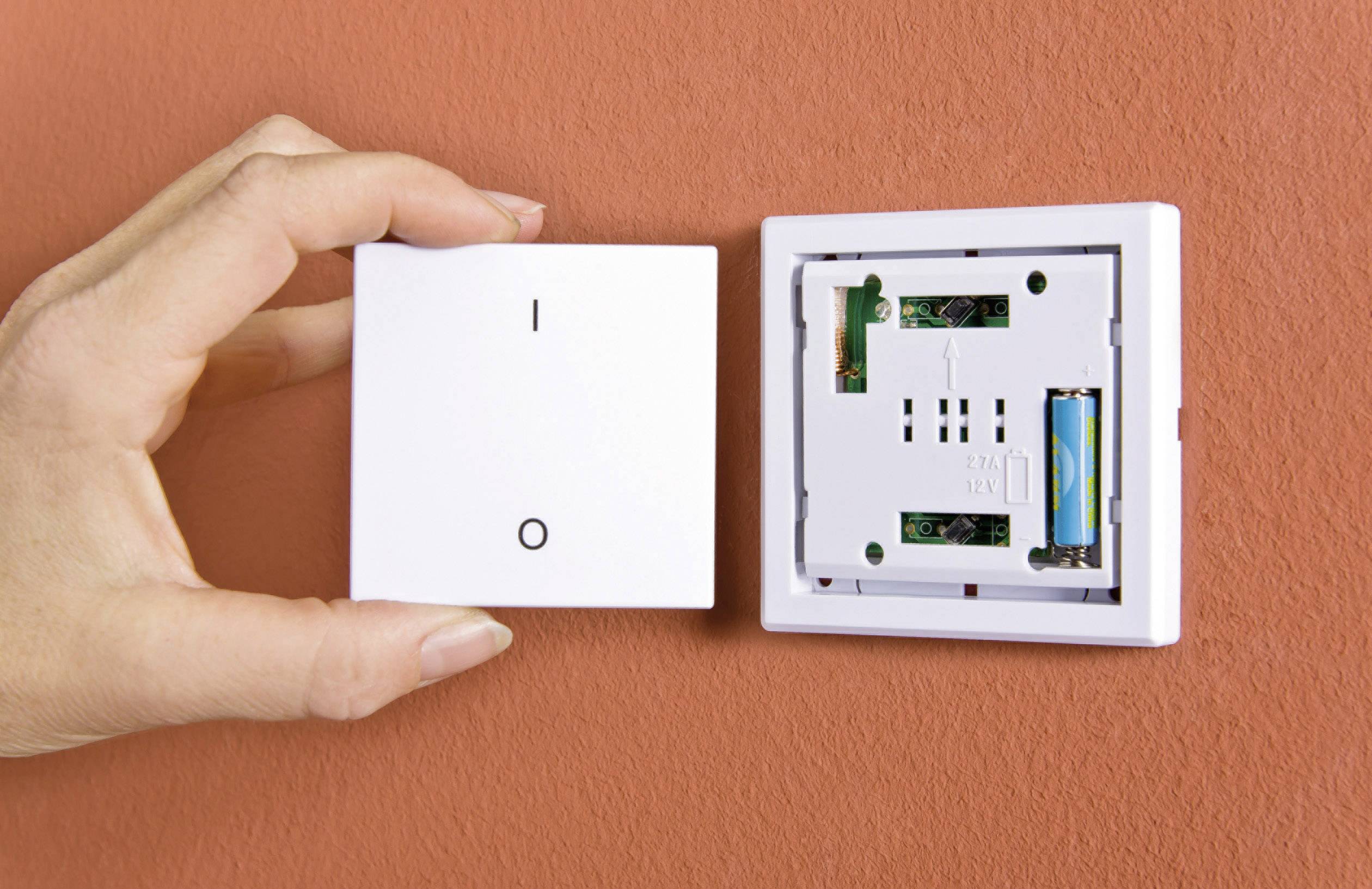 Eine Hand hält die Abdeckung eines Lichtschalters neben der Wand, um ein leeres Gehäuse mit elektronischen Bauteilen zu zeigen.