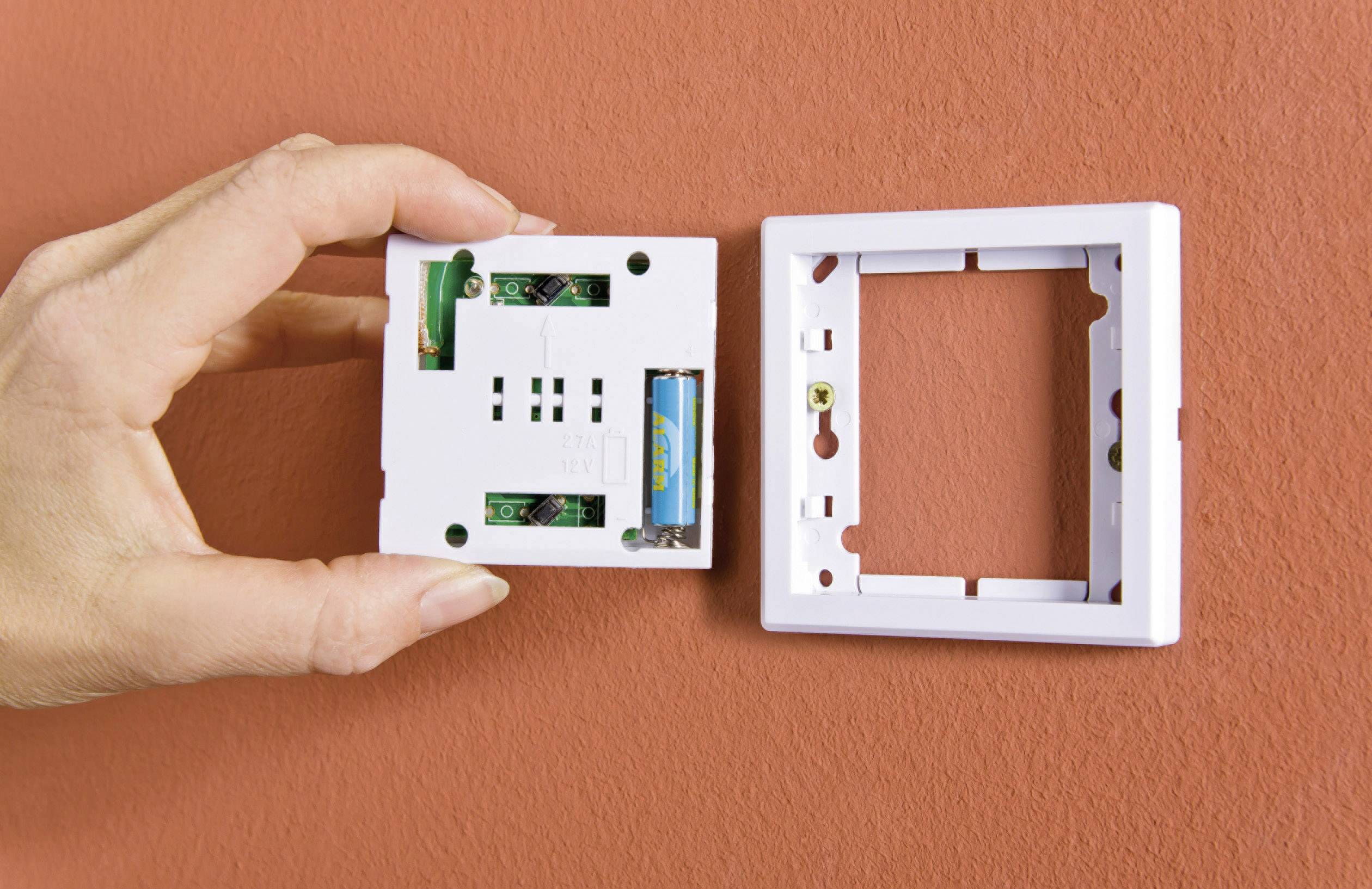 Hand hält einen weißen Lichtschalter ohne Abdeckung, daneben die Abdeckplatte, montiert an einer orangefarbenen Wand.