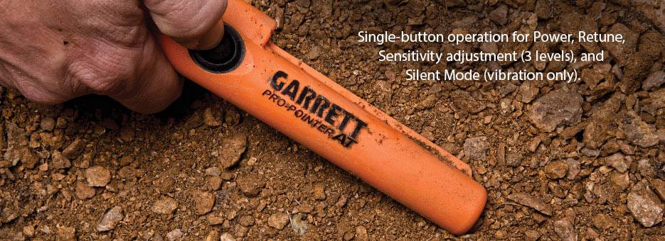 Eine Hand hält einen orangefarbenen Metalldetektor mit der Aufschrift 'GARRETT PRO-POINTER AT'. Text beschreibt Funktionen des Geräts.