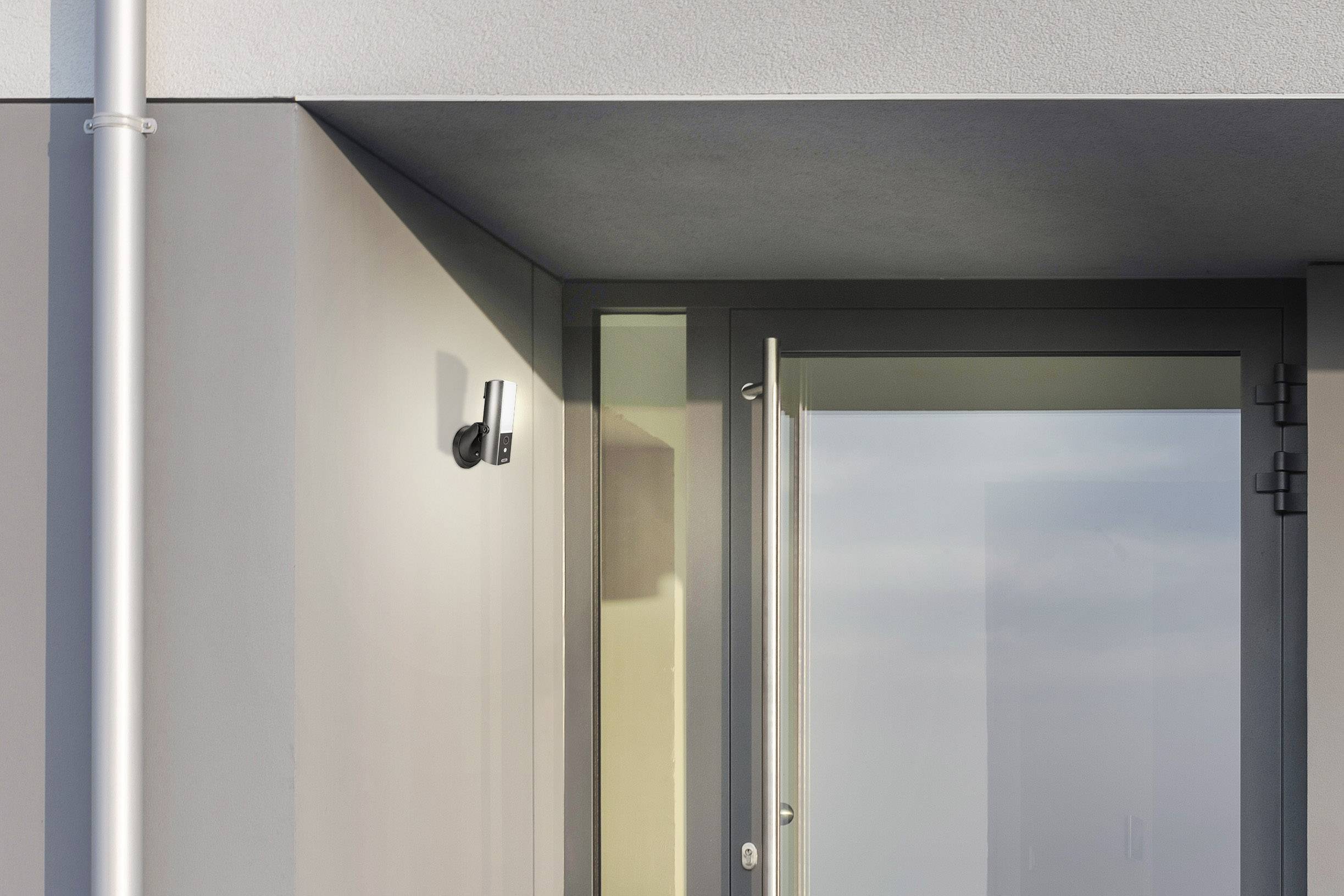 Außenansicht einer modernen Gebäudefassade mit einer kleinen Überwachungskamera neben einer Glastür; klare, minimalistische Architektur.