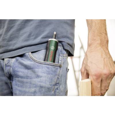 Eine Person in Jeans hält ein Holzstück in der rechten Hand; ein grüner Bosch-Akkuschrauber steckt in der Hosentasche.