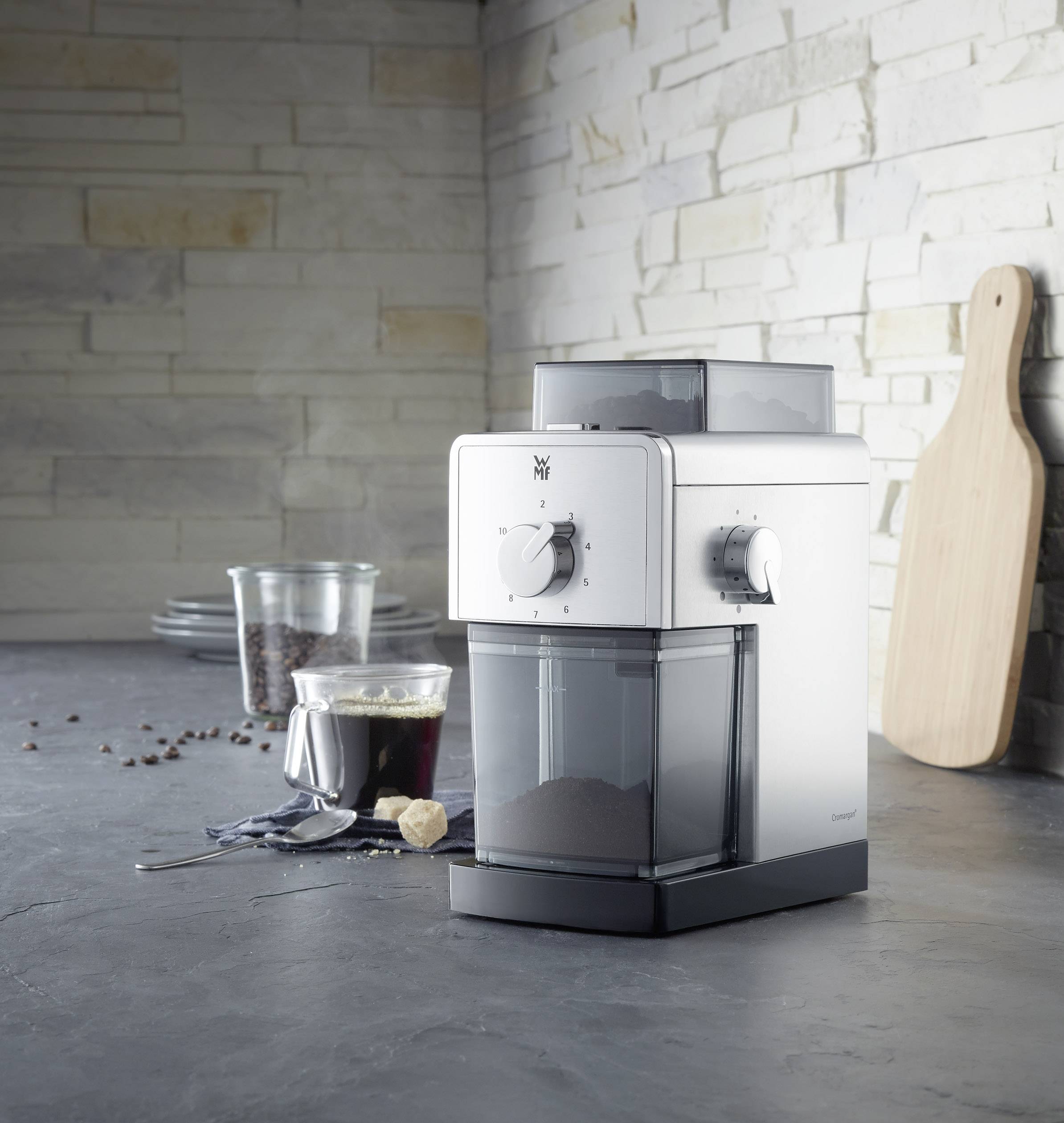 Kaffeemühle auf einer Arbeitsfläche mit Kaffeebohnen, Glas und Brett im Hintergrund. Geeignet für Kaffeeliebhaber zur Zubereitung frischen Kaffees.