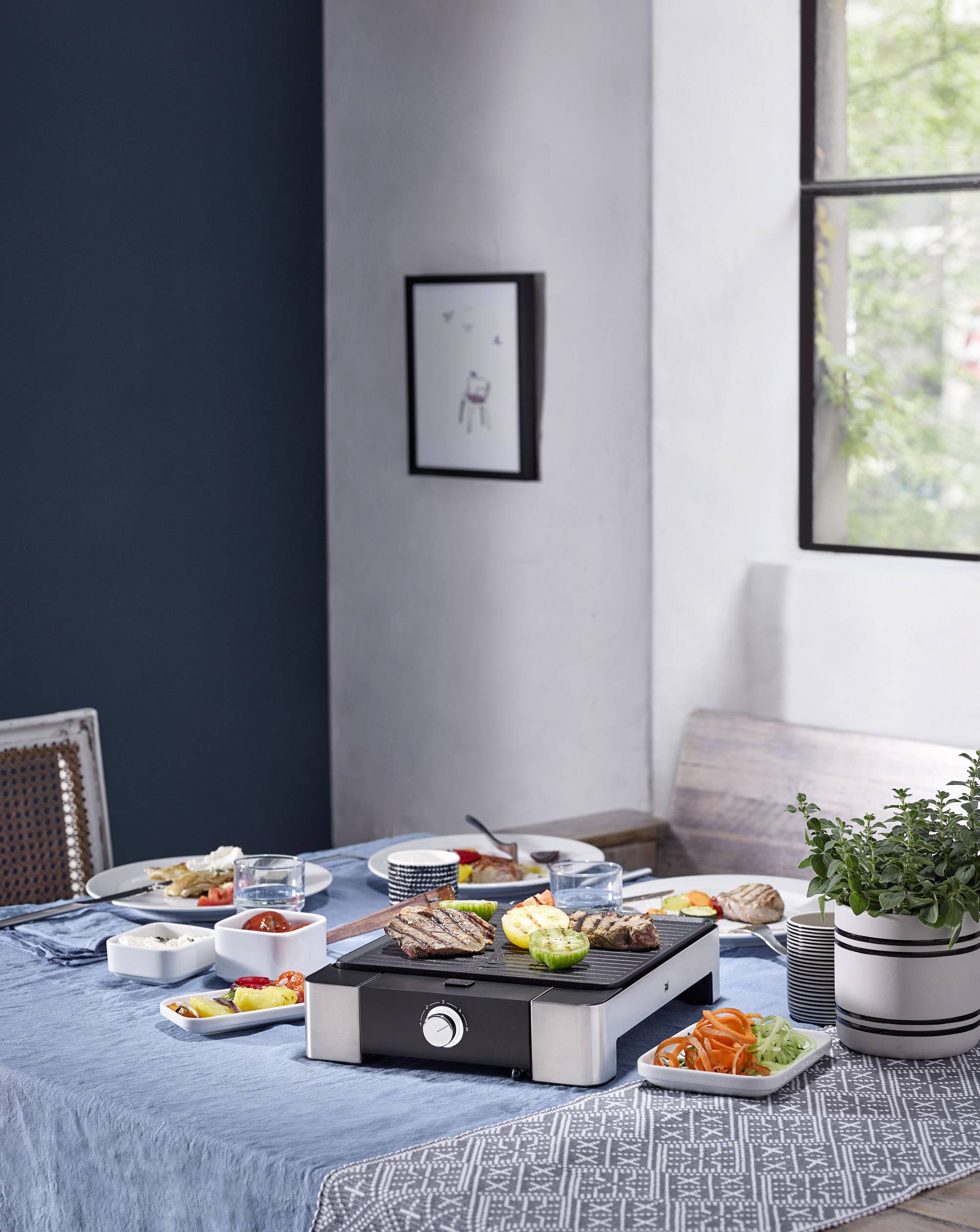 Ein gedeckter Tisch mit einem Elektrogrill in der Mitte, umgeben von Tellern mit Fleisch, Gemüse und Brot. Fenster im Hintergrund.