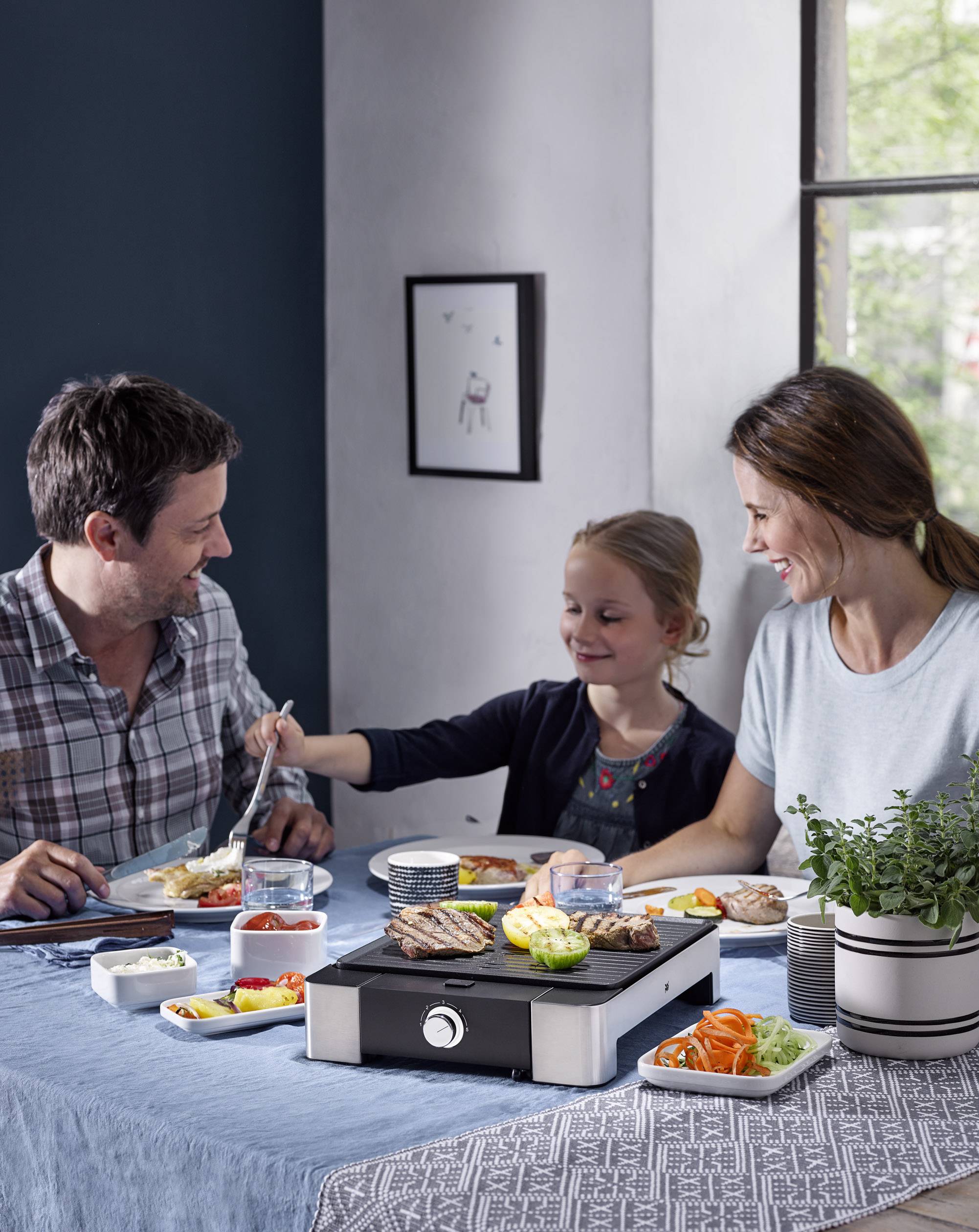 Familie sitzt am Tisch beim Essen, nutzt einen Tischgrill. Ein Mädchen reicht Essen an den Vater, Mutter lächelt. Entspannte Atmosphäre.