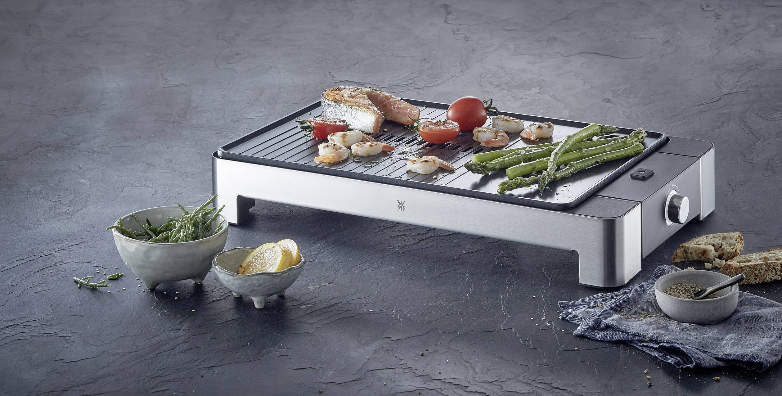 Ein Tischgrill mit Gemüse, Garnelen und Lachs. Daneben stehen Schalen mit Kräutern und Zitrone. Der Hintergrund ist minimalistisch.