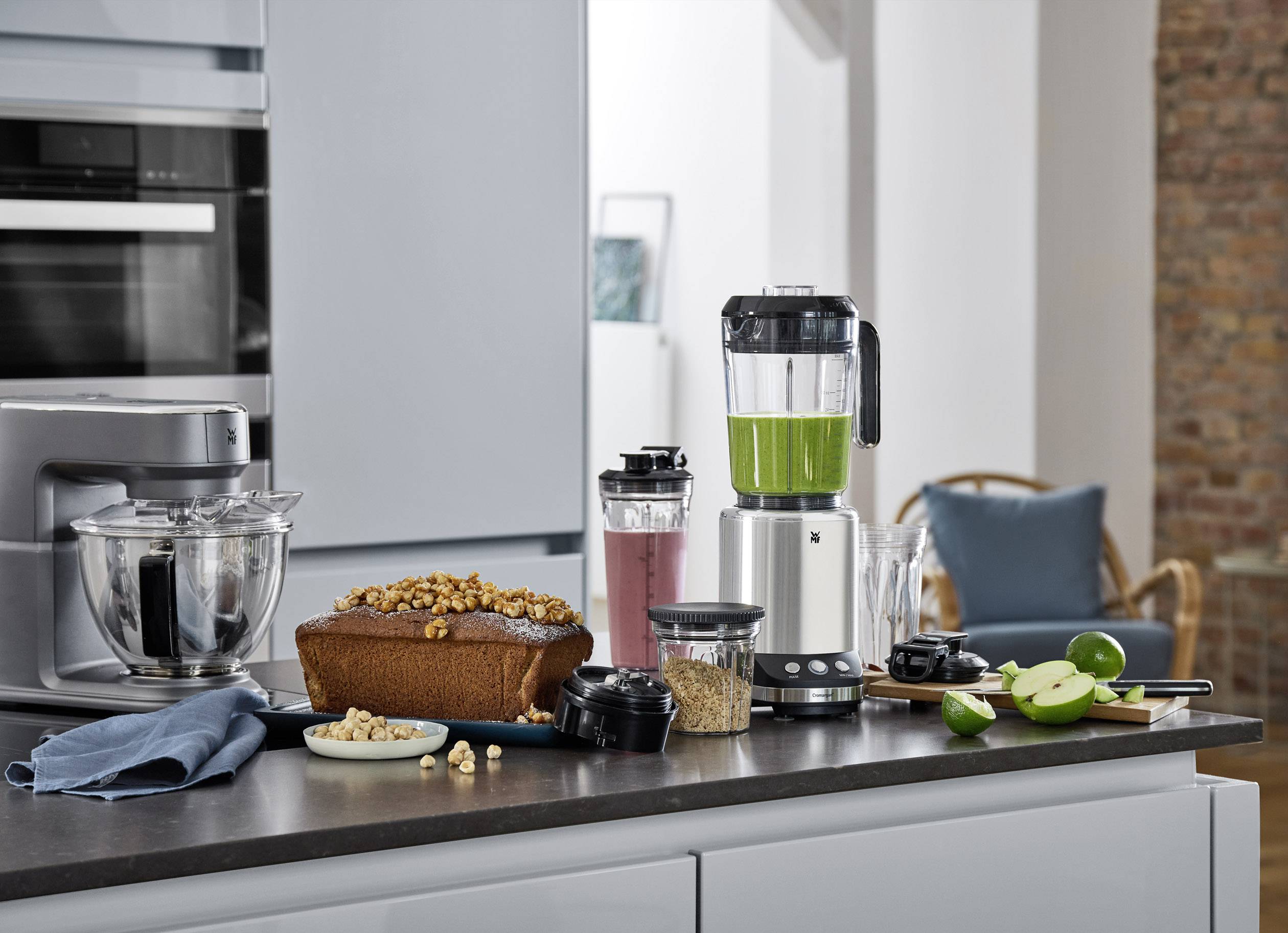 Ein moderner Mixer auf einer Küchenarbeitsplatte mit grünem Smoothie im Behälter. Daneben stehen ein Kuchen, Nüsse und Äpfel.