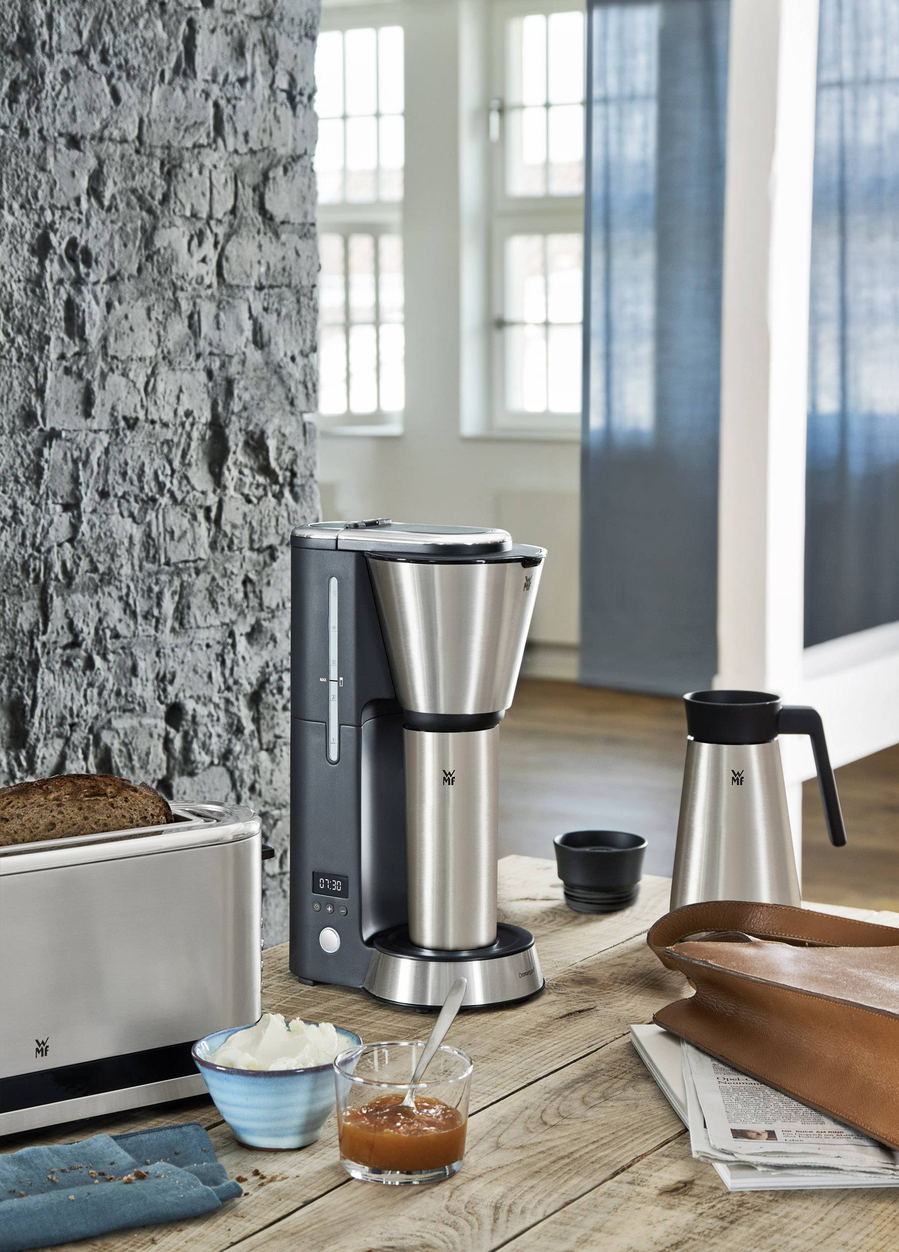 Eine silberne Kaffeemaschine steht auf einem Holztisch neben einem silbernen Toaster, einer Thermoskanne, einem Teller mit Brot und Konfitüre.