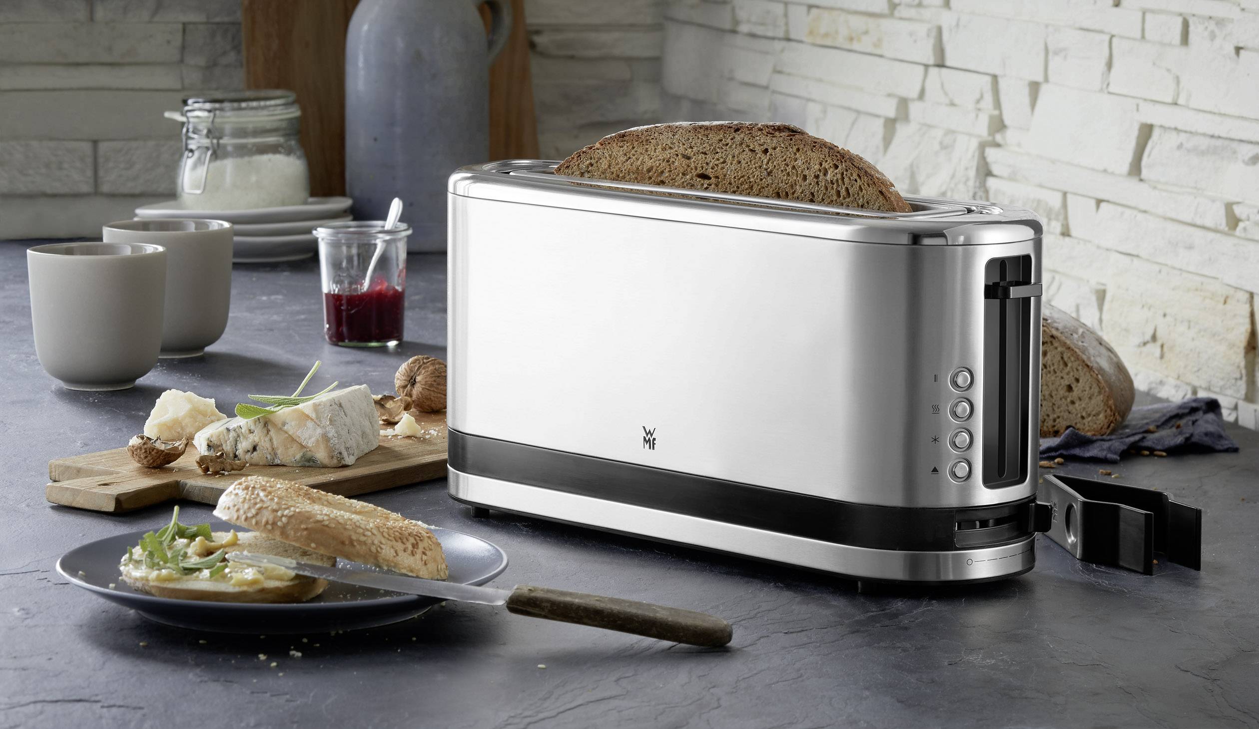 Silberner Toaster mit zwei getoasteten Brotscheiben auf einem Küchentisch, umgeben von Marmelade, Brot und Geschirr.