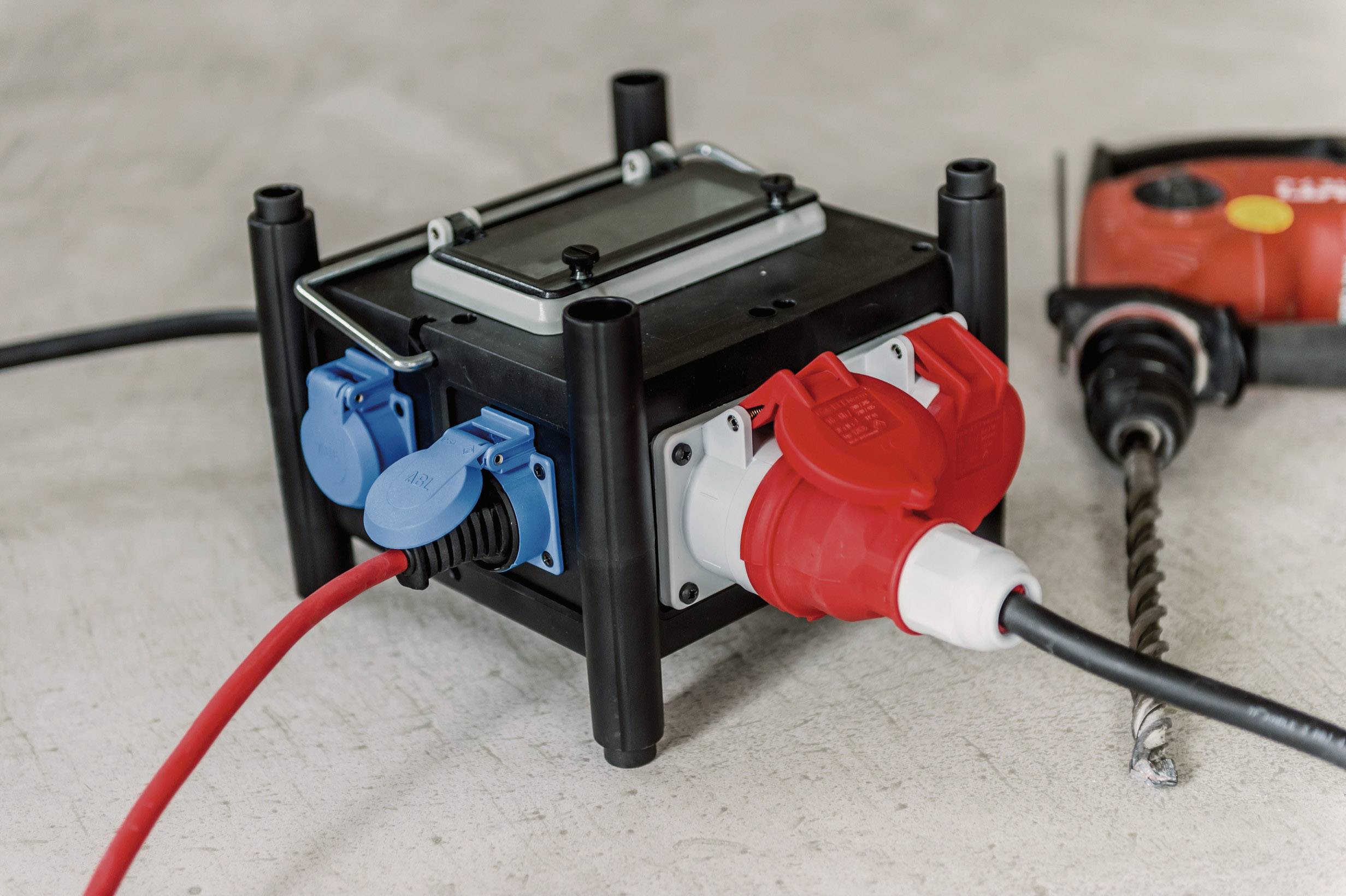 Eine schwarze Steckdosenleiste mit mehreren blauen und roten Anschlüssen, an die ein rotes Kabel angeschlossen ist, neben einer Bohrmaschine.