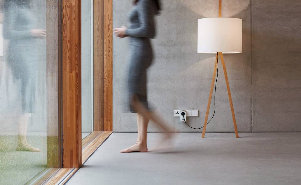 Eine Person geht barfuß in einem Raum mit moderner Einrichtung und großer Glasfront. Daneben steht eine Lampe mit Holzstativ.