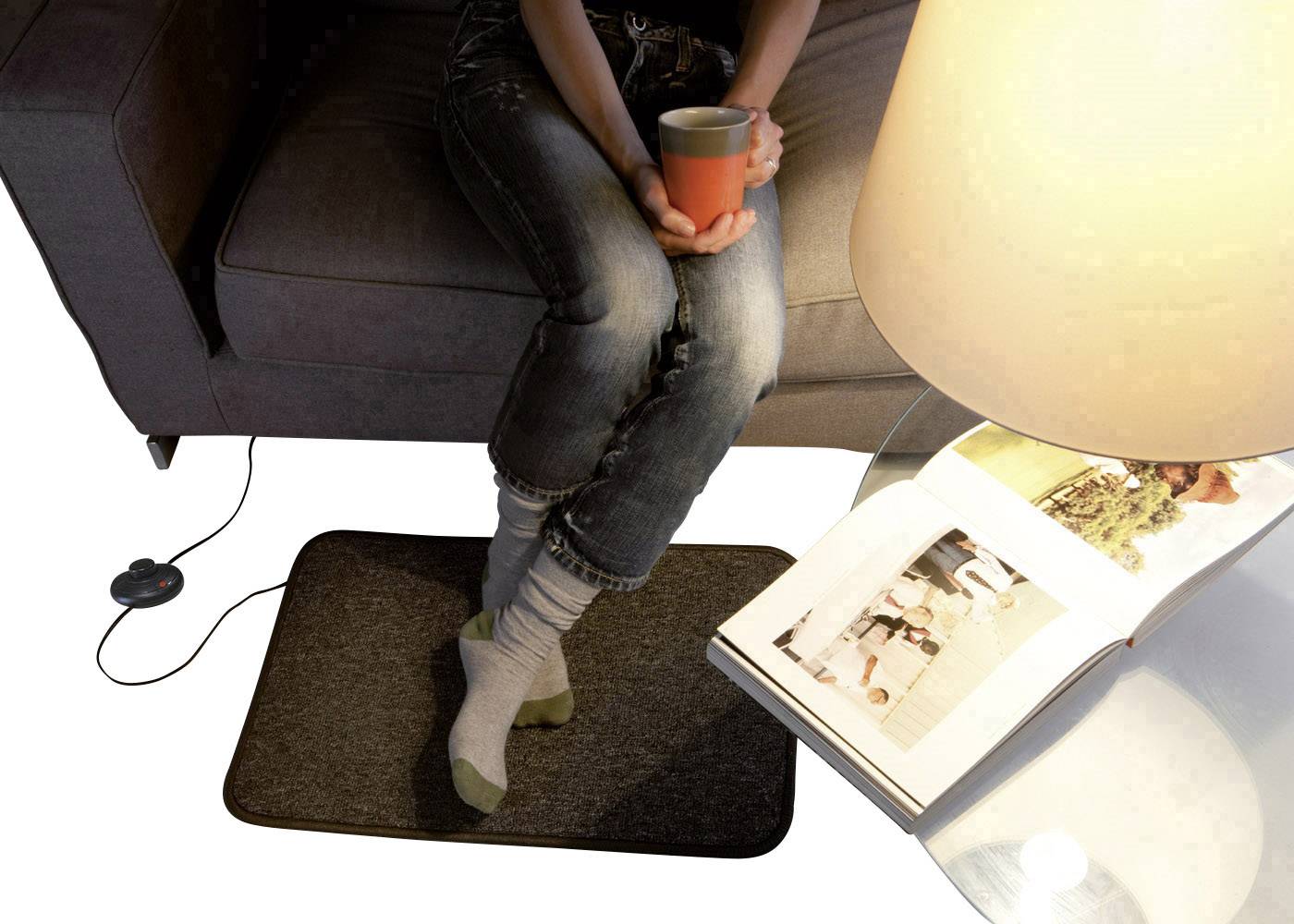 Person auf Sofa hält Tasse, Füße auf beheiztem Teppich. Nebenan Lampe und aufgeschlagenes Buch auf Tisch. Wohnzimmeratmosphäre.