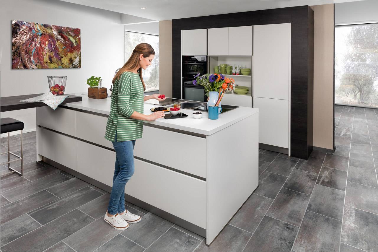 Eine Frau steht in einer modernen Küche an einer weißen Insel und schneidet Obst. Im Hintergrund sind Schränke und Dekorationen zu sehen.