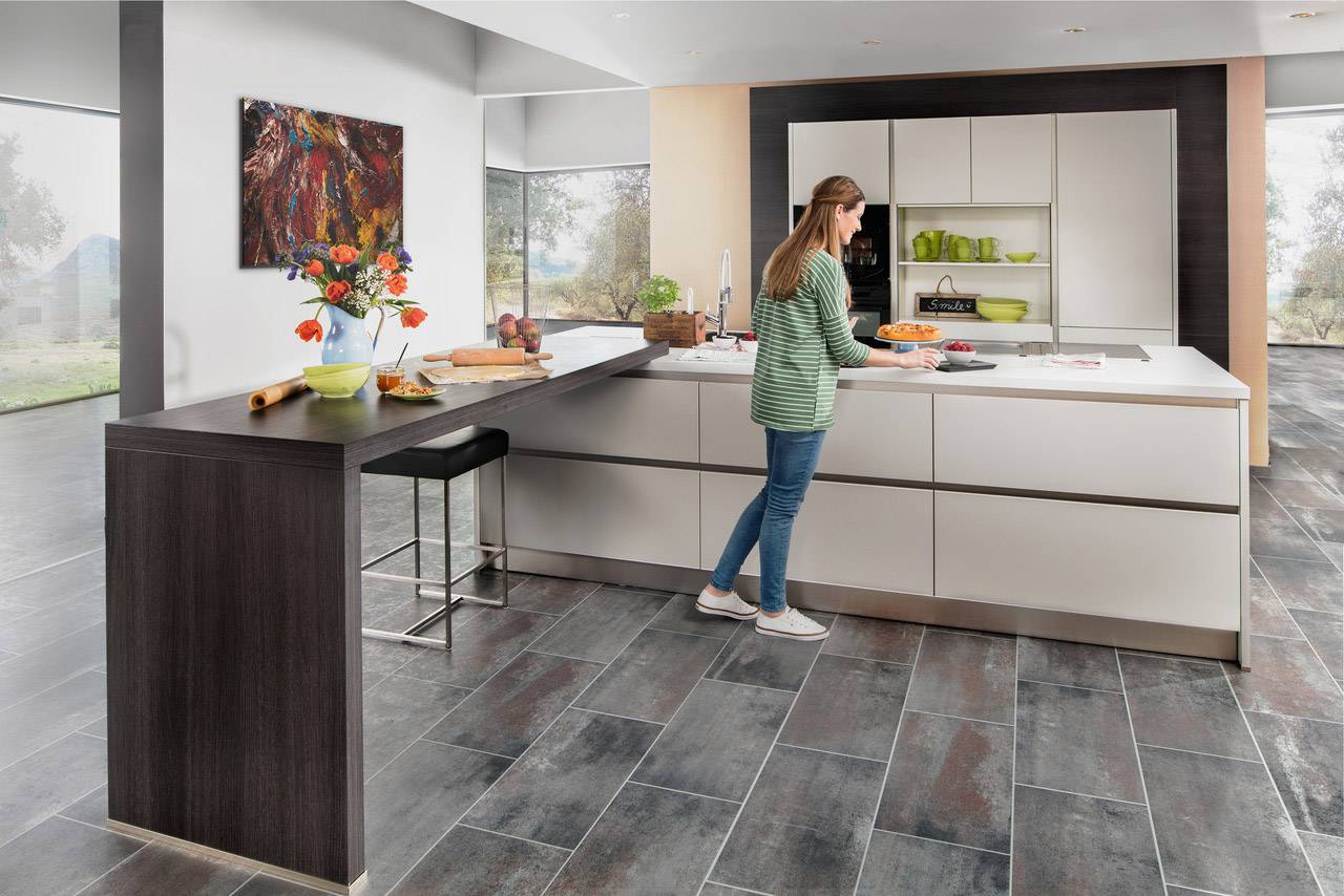 Eine Frau steht in einer modernen Küche mit grauem Fliesenboden und bereitet Essen auf der weißen Arbeitsplatte zu. Im Hintergrund sind ein großes Fenster und ein Kunstwerk an der Wand zu sehen.