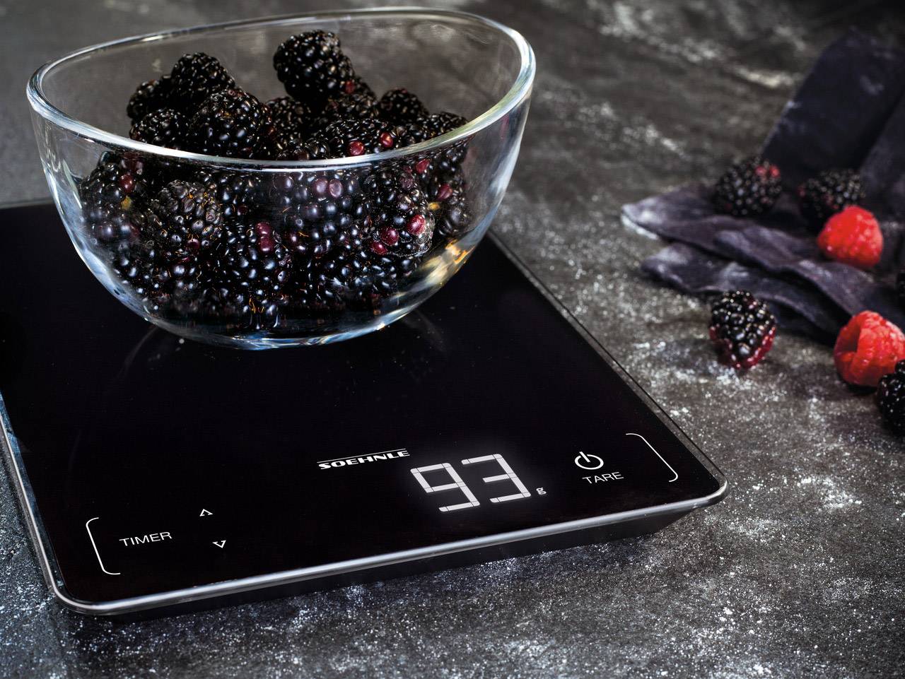 Eine Glasschale voll mit Brombeeren steht auf einer digitalen Küchenwaage, die 93 Gramm anzeigt. Daneben liegen weitere Beeren auf einer dunklen Oberfläche.