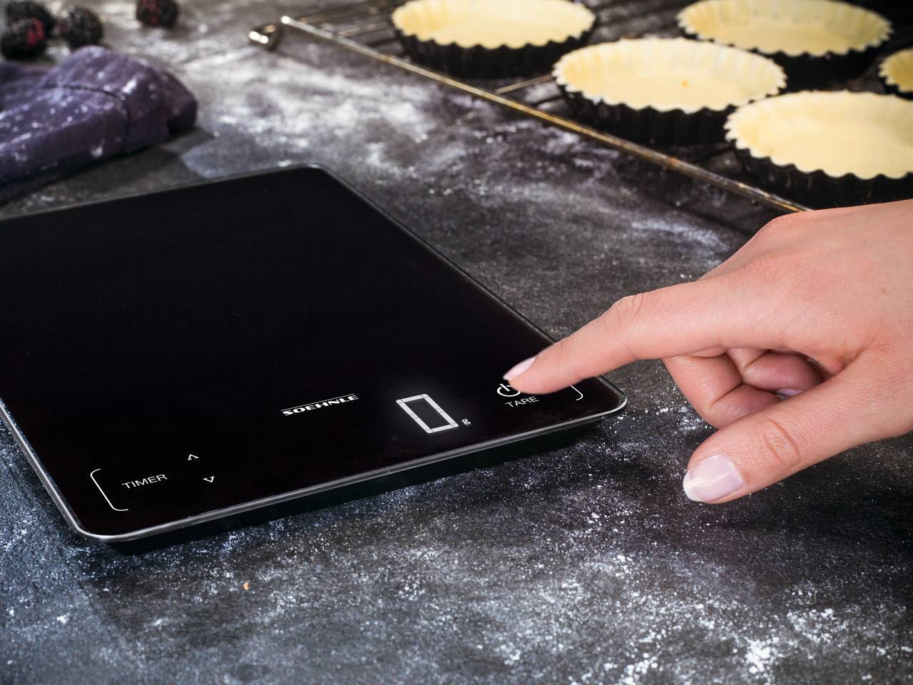 Eine Hand bedient eine digitale Küchenwaage auf einer bemehlten Arbeitsfläche, daneben sind ungebackene Tartes in Backformen.