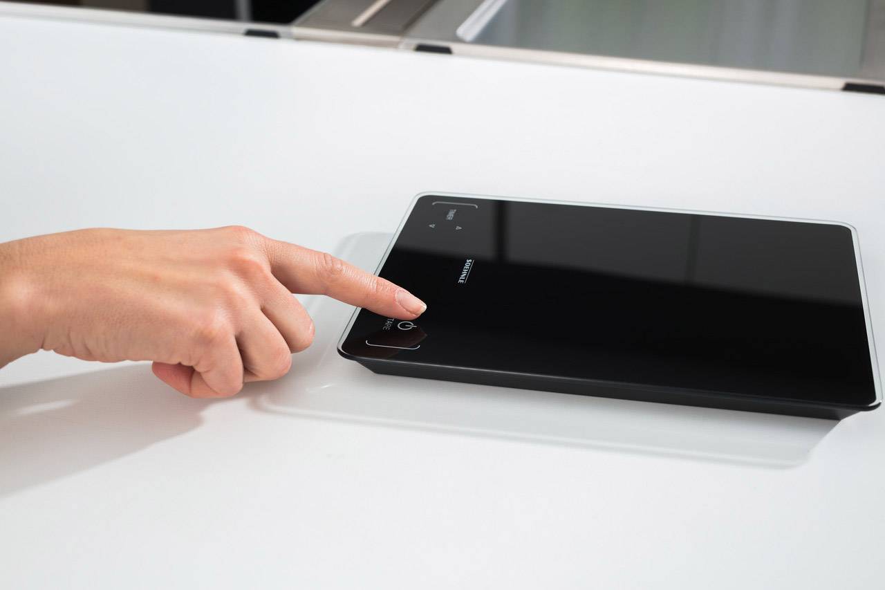 Eine Hand berührt ein digitales Gerät mit dunklem Bildschirm; möglicherweise ein Tablet oder Displaygerät auf einem Tisch.
