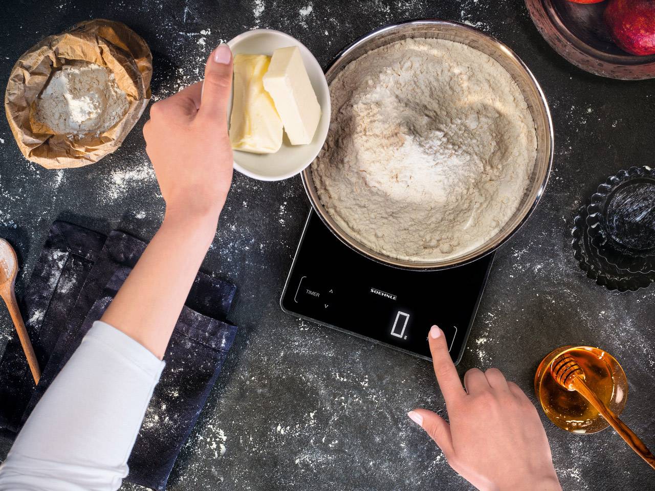Eine Person wiegt Butter auf einer digitalen Küchenwaage neben einer Schüssel mit Mehl, Zucker und einer Tüte Mehl auf einem dunklen Tisch.