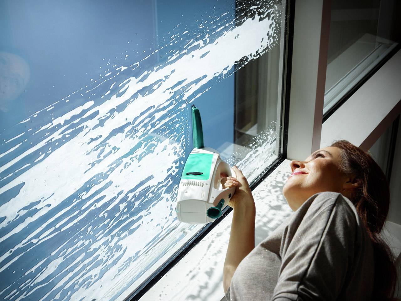 Eine Frau reinigt ein Fenster mit einem elektrischen Fenstersauger. Sie schaut zufrieden, während sie das Gerät über das Glas führt.