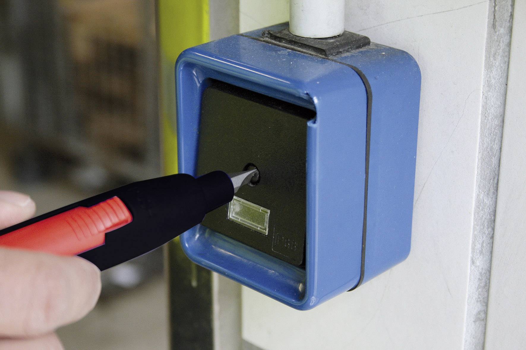 Person hält einen roten Stift an einen blauen Kasten mit kleinem Loch. Der Kasten scheint Teil eines Schließsystems zu sein.