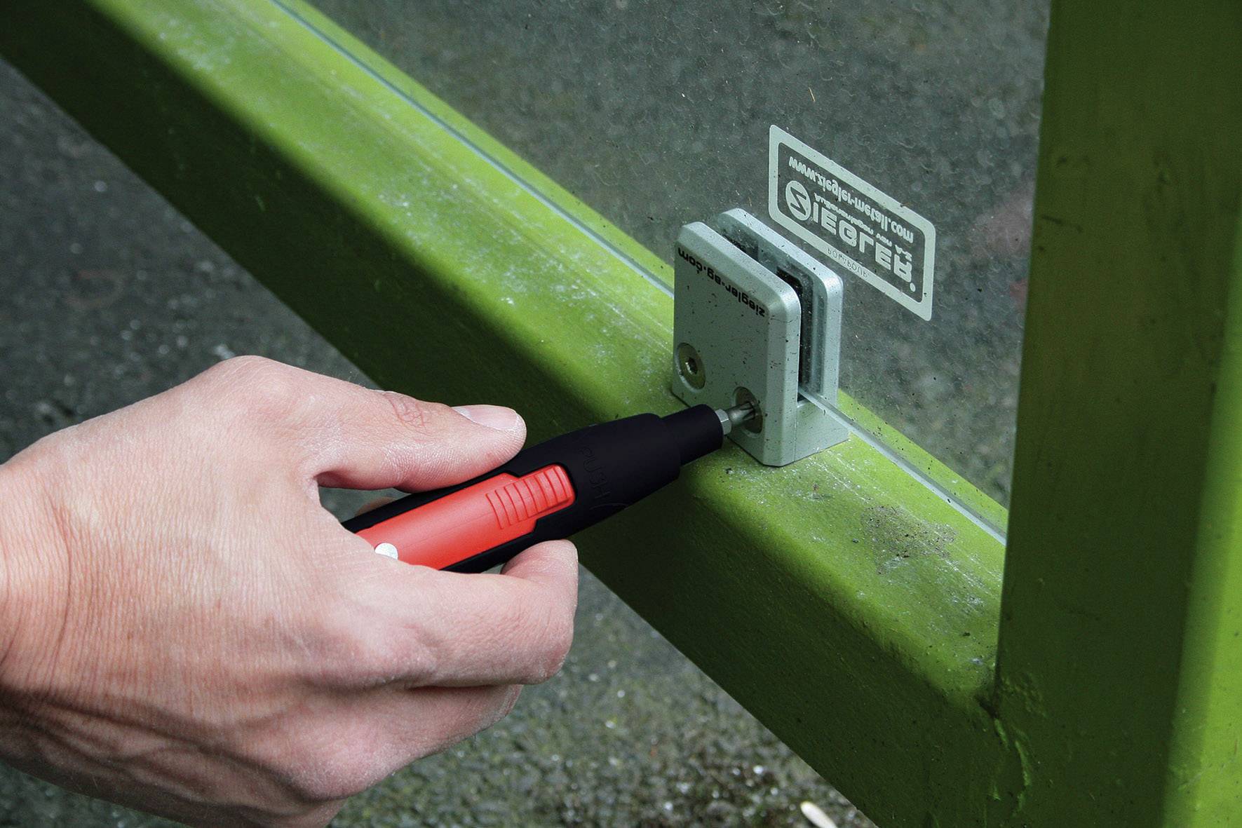 Eine Hand hält einen roten Stift gegen ein Metallscharnier an einem grünen Rahmen.