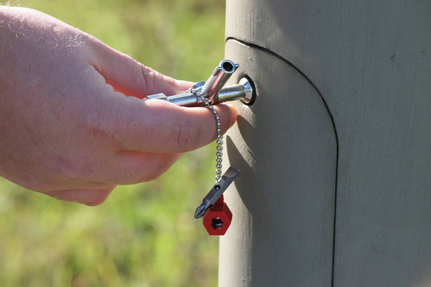 Eine Hand dreht einen Vierkantschlüssel im Schloss einer grauen Säule im Freien. Ein rotes Sechskantwerkzeug hängt am Schlüssel.