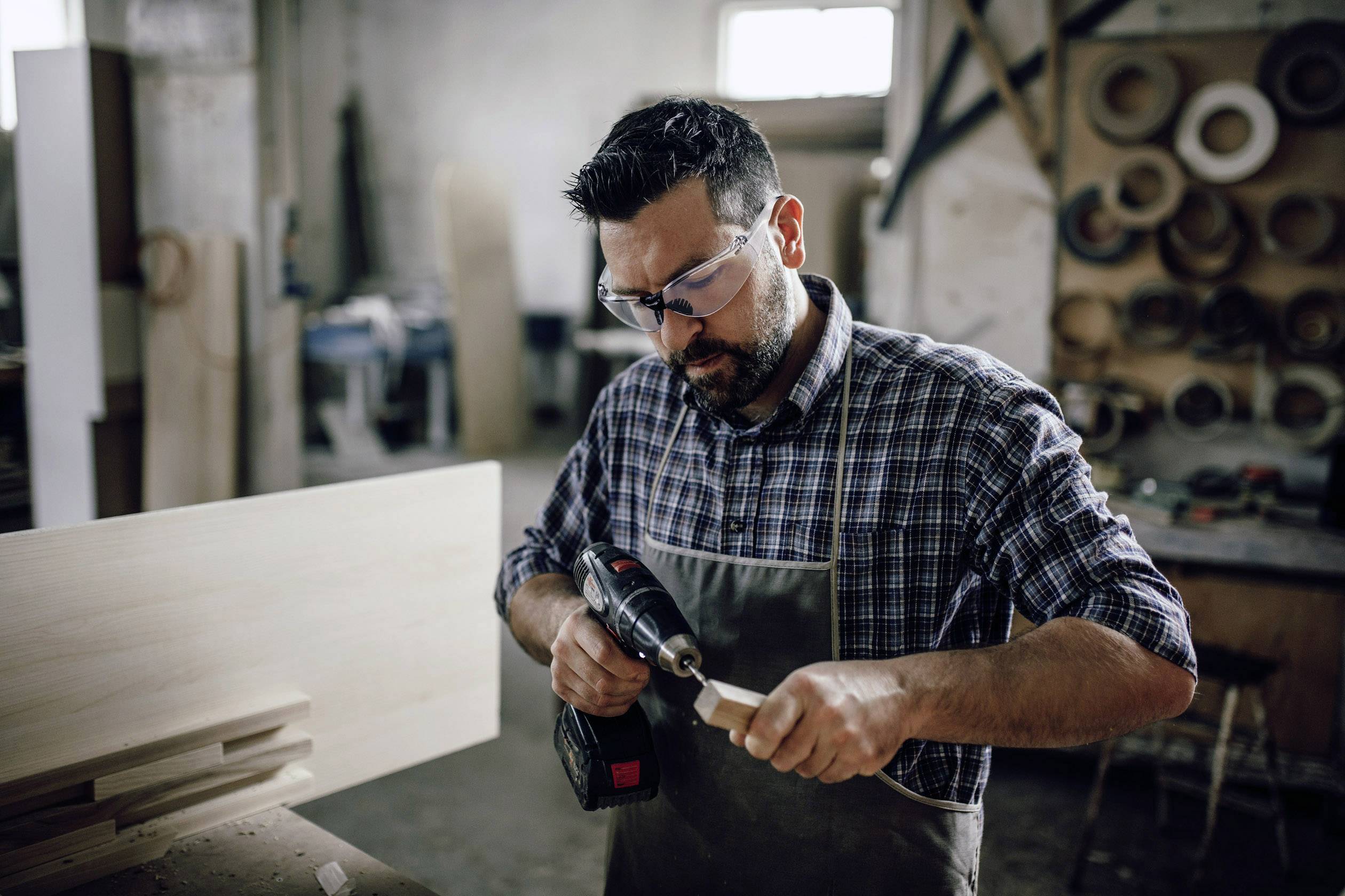 Ein Mann in Schutzbrille und Schürze arbeitet konzentriert mit einem Akkuschrauber in einer Holzwerkstatt.