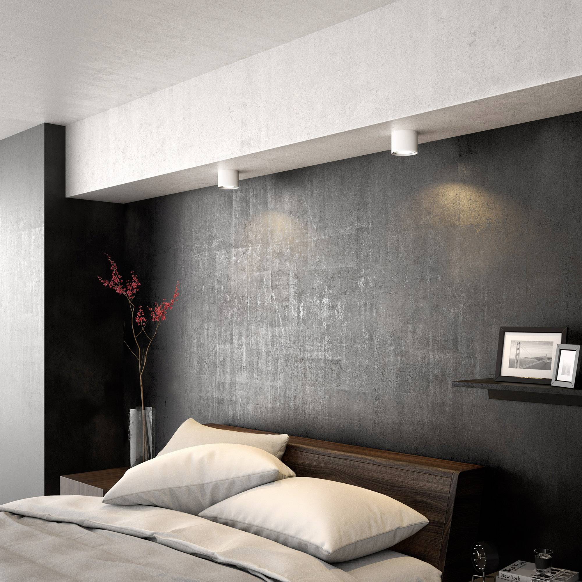 Modernes Schlafzimmer mit einem großen Bett, zwei Kissen und einem gemusterten Bettkissen. An der Wand steht eine hohe Vase mit roten Zweigen.