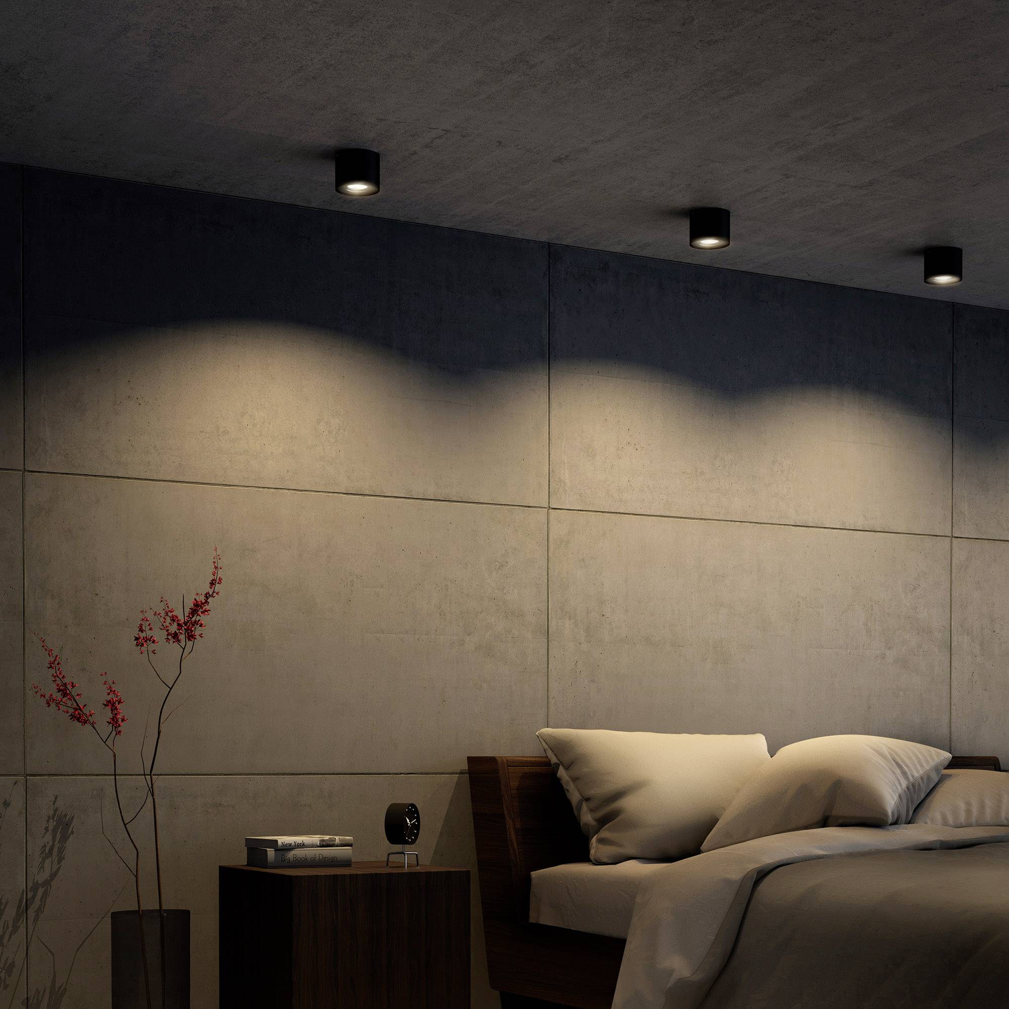 Ein modernes Schlafzimmer mit Betonwänden, gedämpfter Beleuchtung und einem großen Bett mit vielen Kissen. Links steht eine Vase mit Ästen.