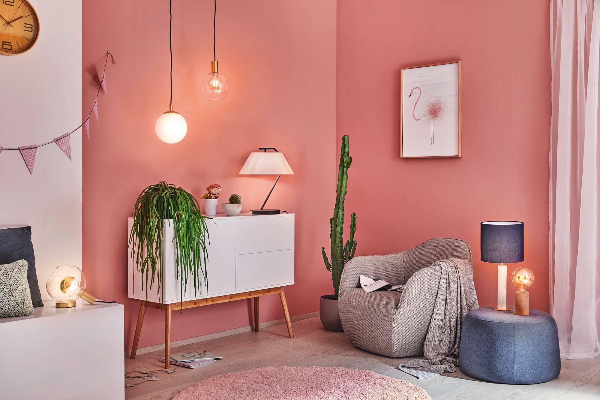 Ein gemütliches Wohnzimmer mit rosa Wänden, modernem Sessel, Kaktus, Hängelampen, einem kleinen Schrank und gerahmtem Bild an der Wand.