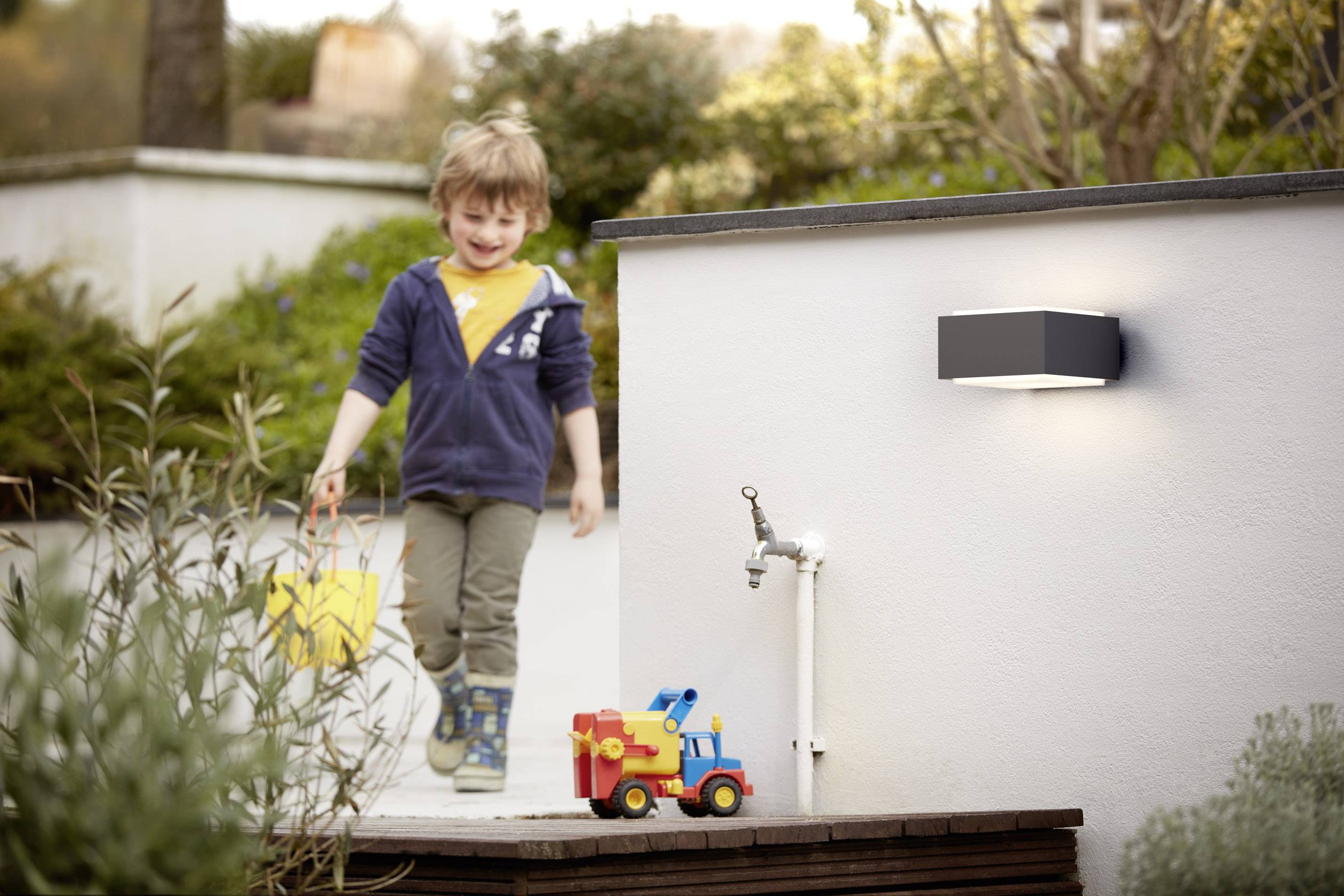 Ein Kind mit einem gelben Eimer geht im Garten. Vor ihm steht ein buntes Spielzeugauto. Eine Wandlampe leuchtet im Hintergrund.