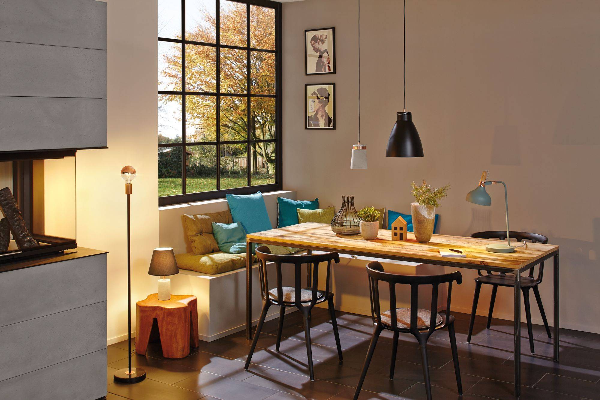 Esszimmer mit Holztisch, vier Stühlen und zwei Lampen. Ein großes Fenster zeigt herbstliche Bäume. Kissenbank unter dem Fenster.