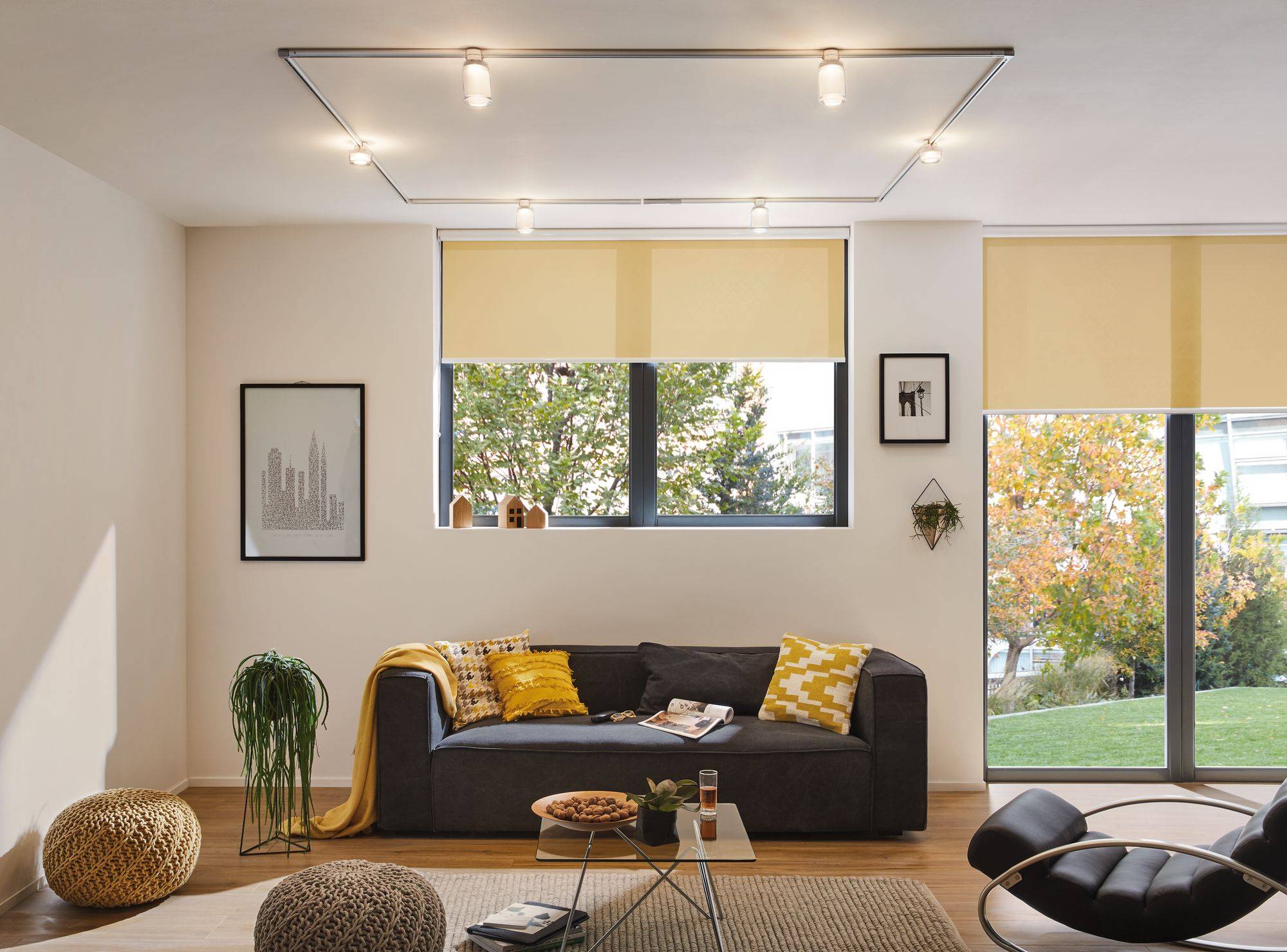 Modernes Wohnzimmer mit grauem Sofa, das mit gelben Kissen dekoriert ist. Große Fenster mit Rollos bieten Blick auf grüne Bäume.