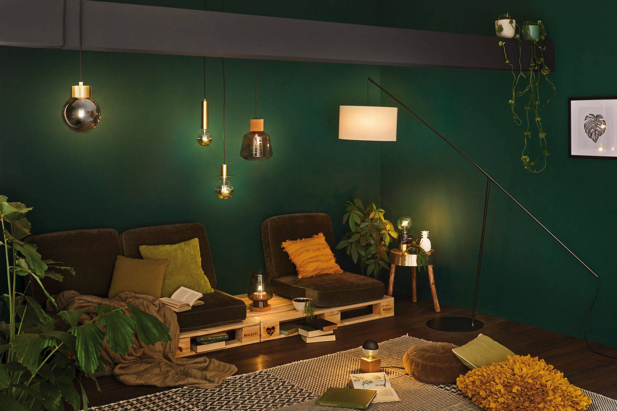Ein gemütlicher Wohnbereich mit grünen Wänden, Palettensofa, Kissen und Decken. Verschiedene Lampen beleuchten den Raum. Pflanzen und Bücher schaffen eine entspannte Atmosphäre.