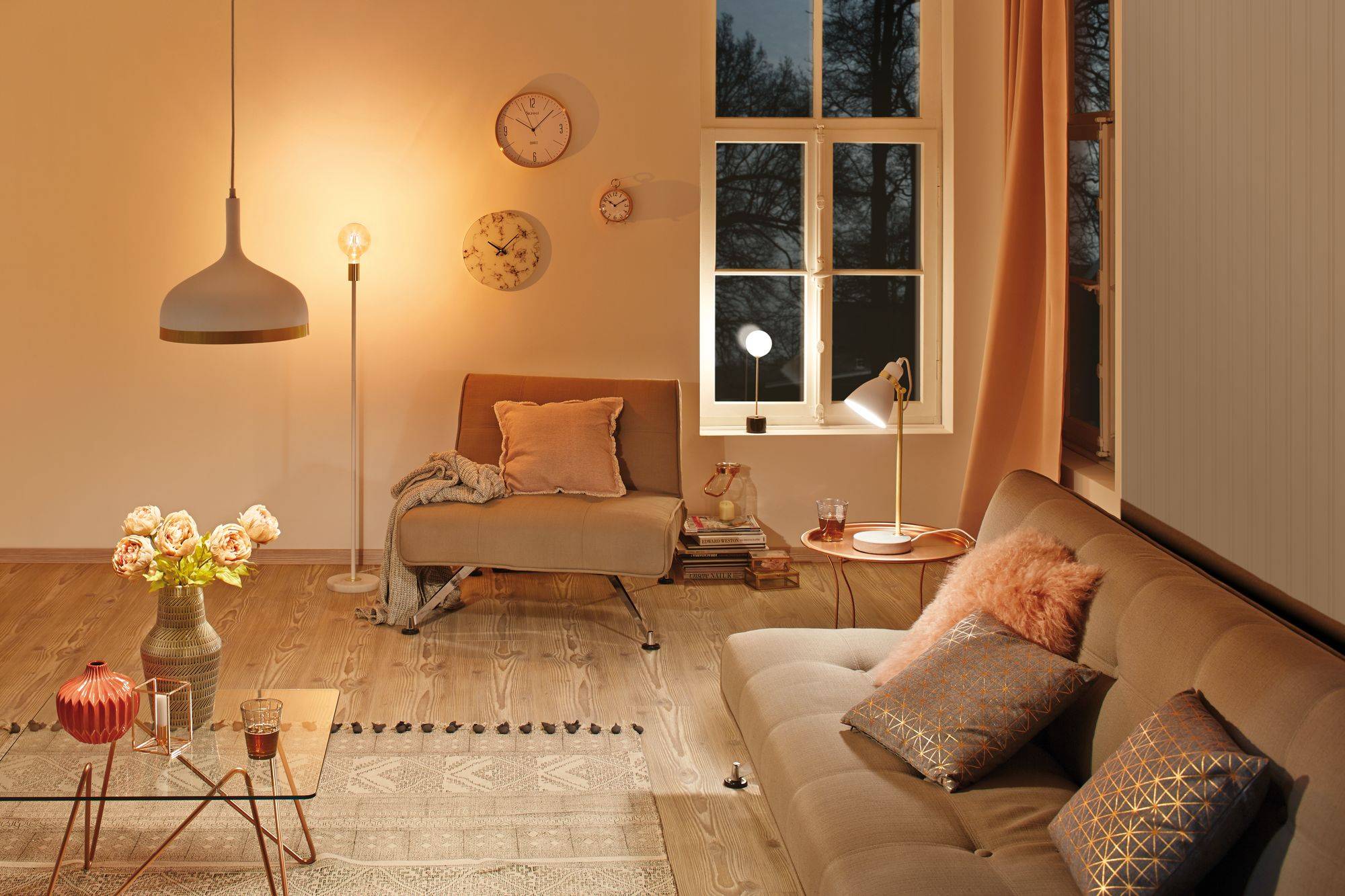 Gemütliches Wohnzimmer mit brauner Couch, Kissen, Stehlampe und verglastem Tisch. Warme Beleuchtung und Fenster im Hintergrund.
