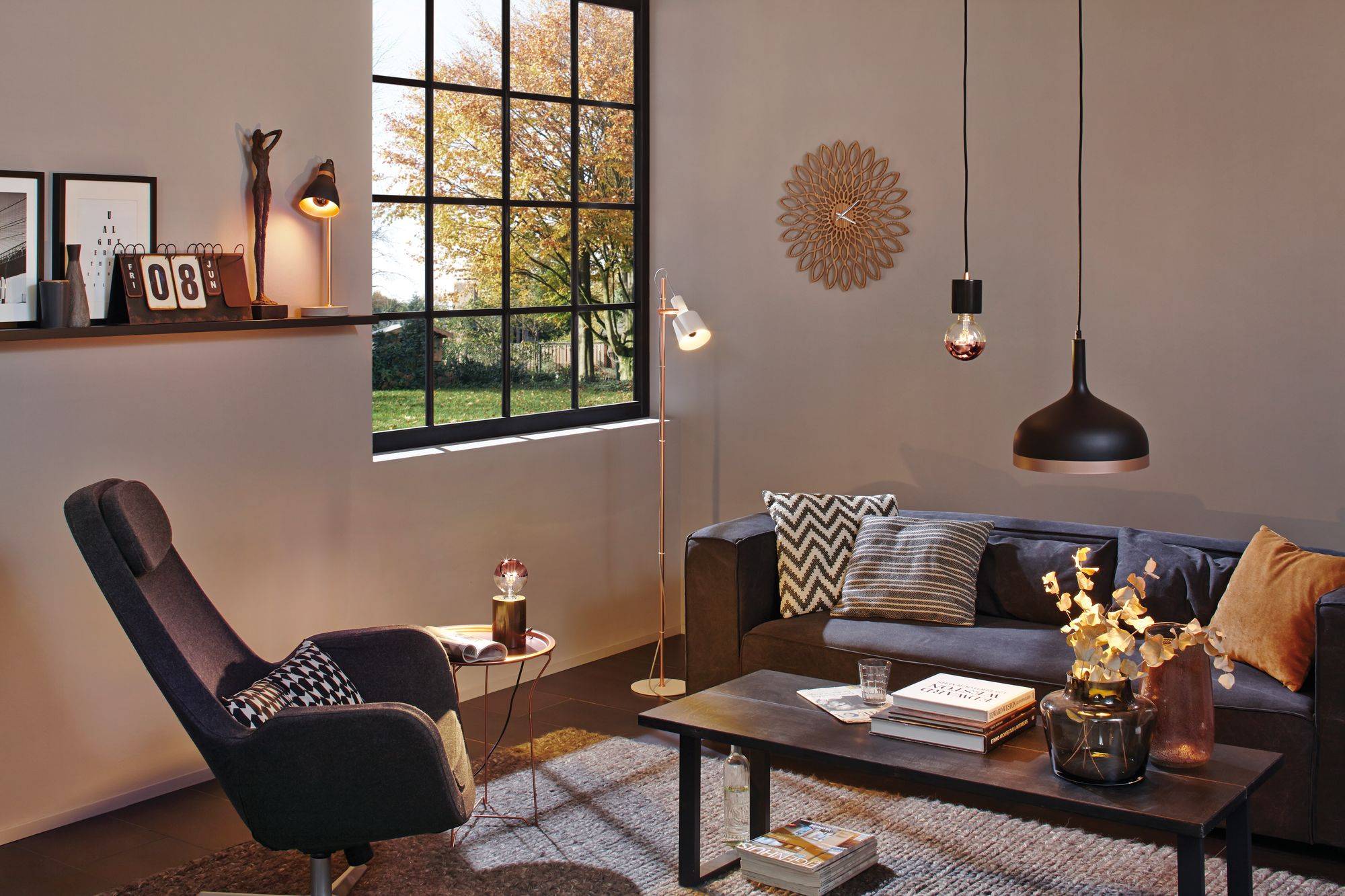 Gemütliches Wohnzimmer mit Sessel, Sofa und Couchtisch. Große Fenster öffnen den Raum zum herbstlichen Außenbereich. Warmes Licht schafft eine einladende Atmosphäre.