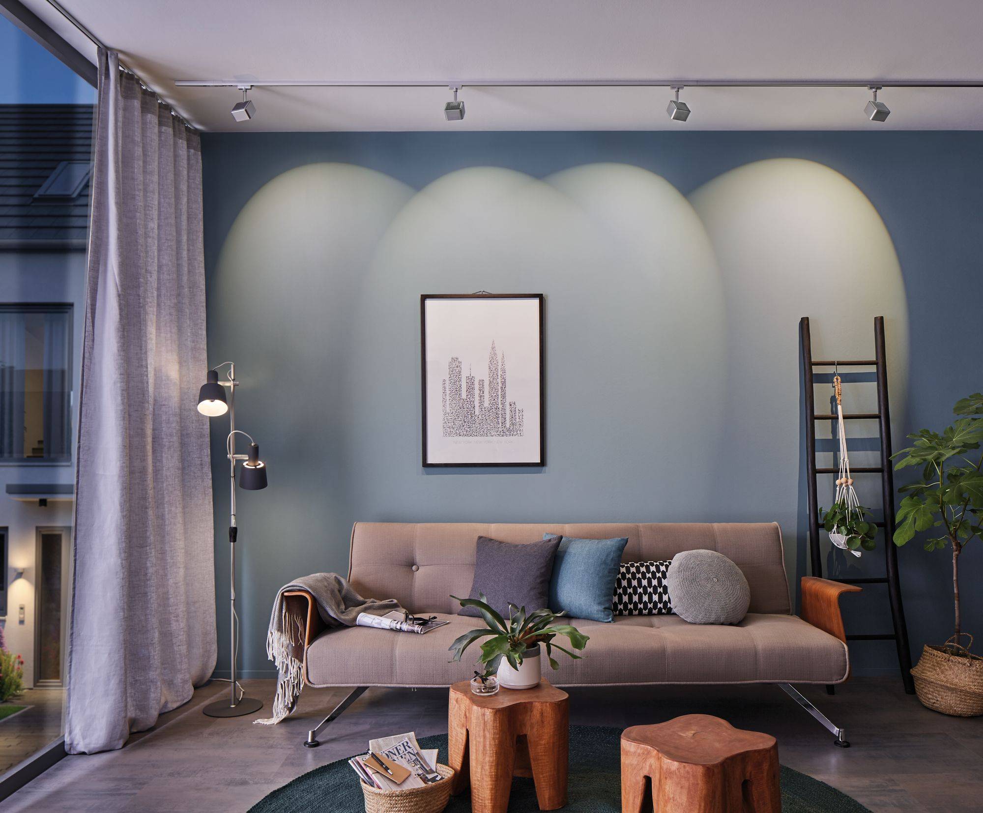 Wohnzimmer mit blau gestrichener Wand, beige Sofa, runden Holztischen und Pflanzen. Ein gerahmtes Stadtbild hängt an der Wand.