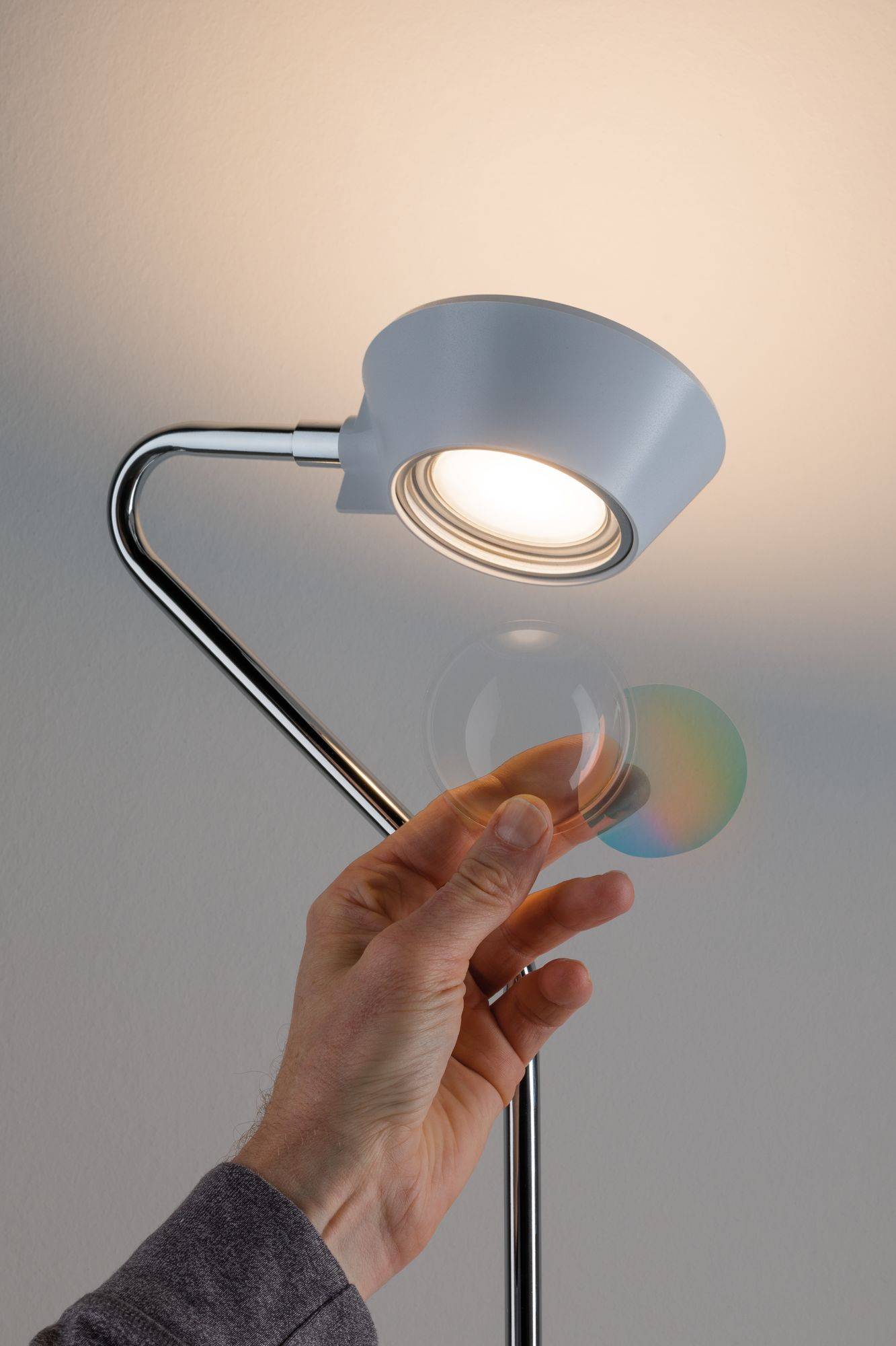 Eine Hand hält eine Linse vor eine weiße Lampe, wodurch ein bunter Lichtschein auf der Wand erzeugt wird.