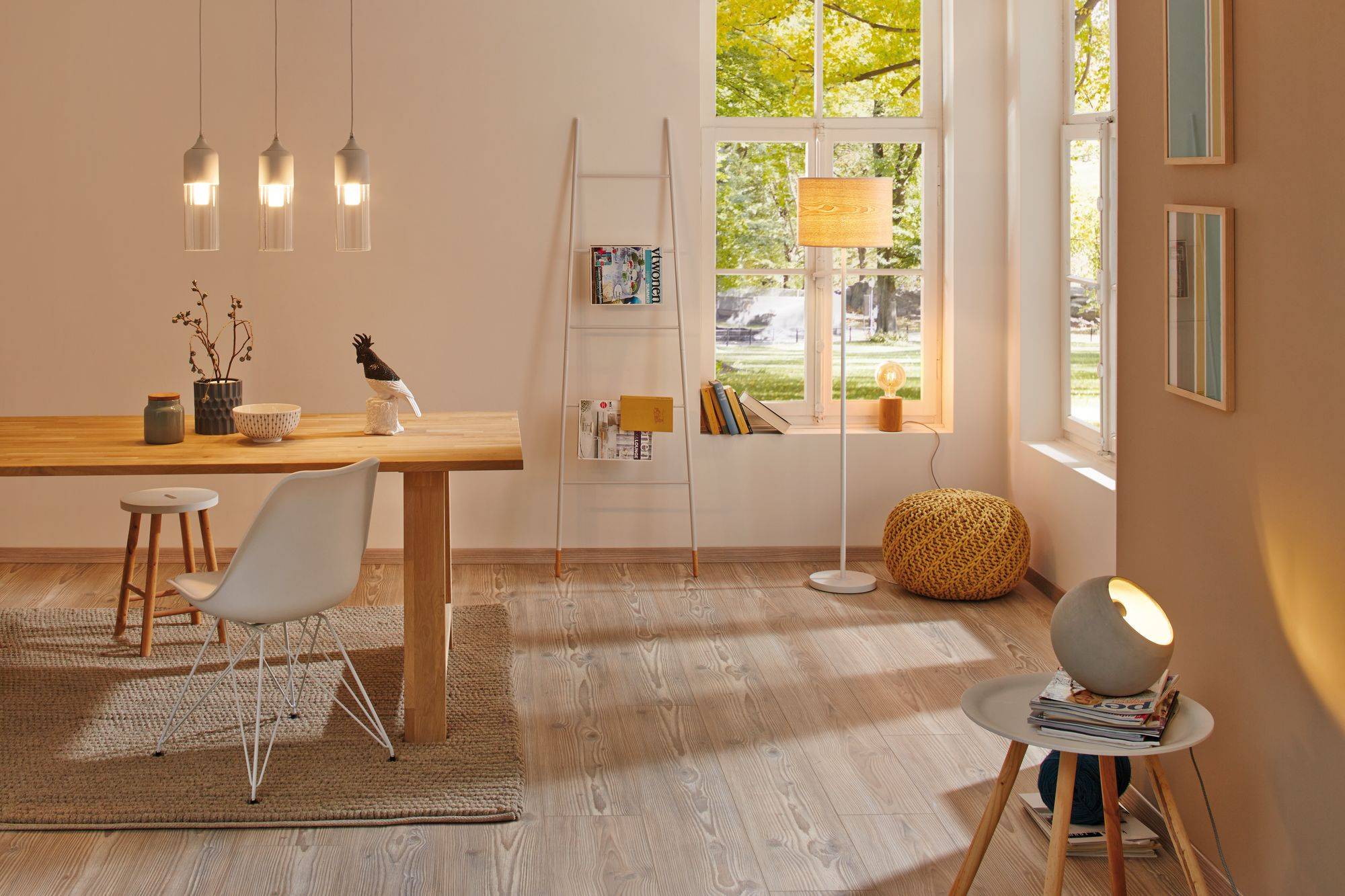 Ein helles Wohnzimmer mit großem Holztisch, weißen und holzfarbenen Stühlen. Stehlampe und Fenster mit Blick auf grüne Bäume.