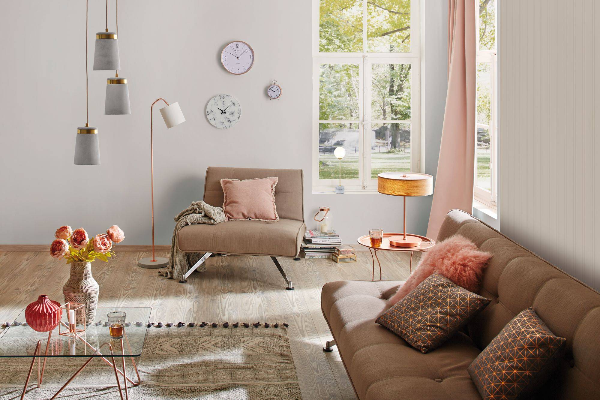 Ein Wohnzimmer mit Sofa und Sessel in neutralen Farben, dekoriert mit Uhren und Leuchten. Große Fenster lassen Tageslicht herein.