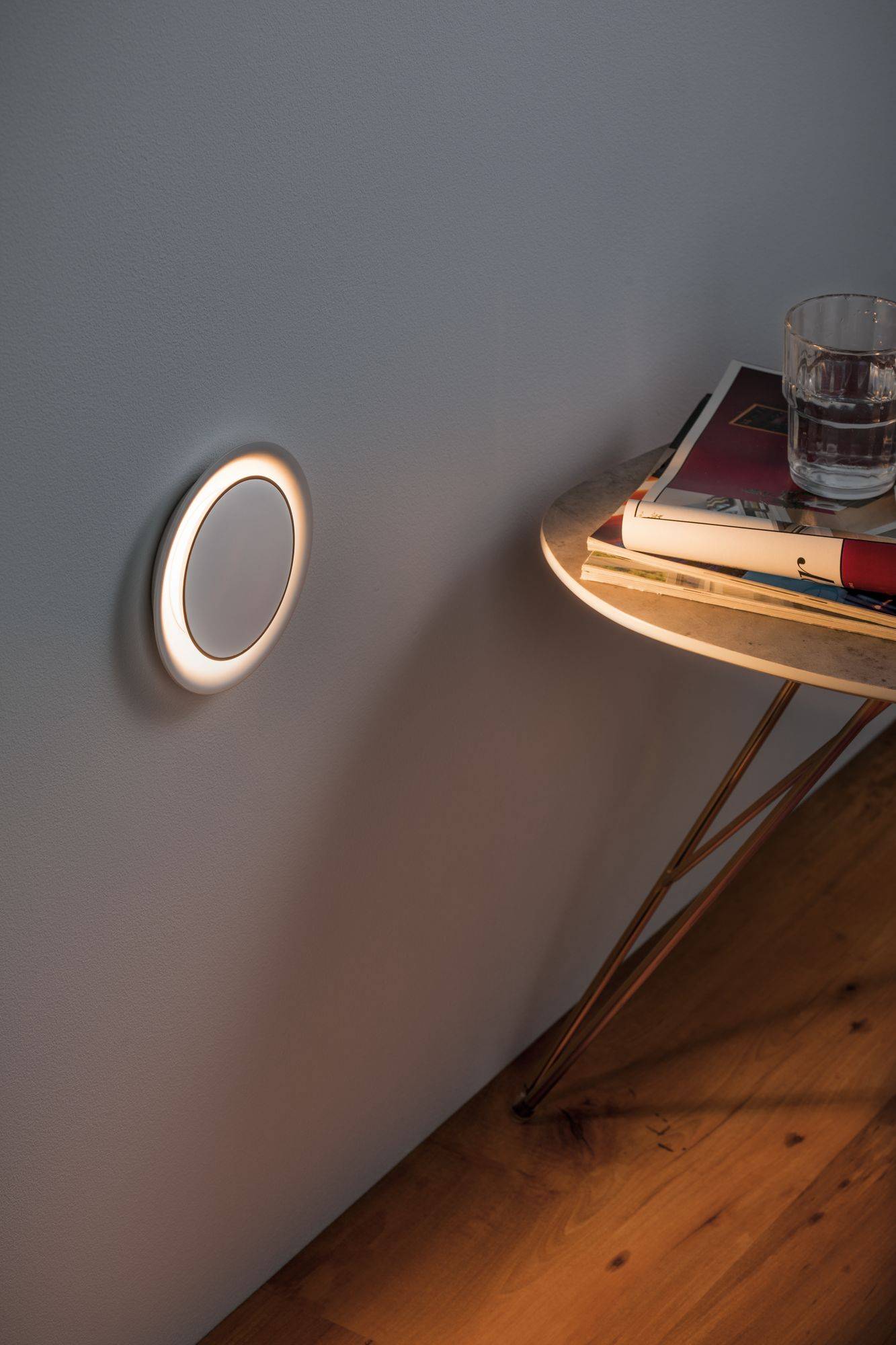 Eine runde Lampe leuchtet an der Wand. Daneben steht ein dreibeiniger Tisch mit Büchern und einem Glas Wasser auf Holzfußboden.