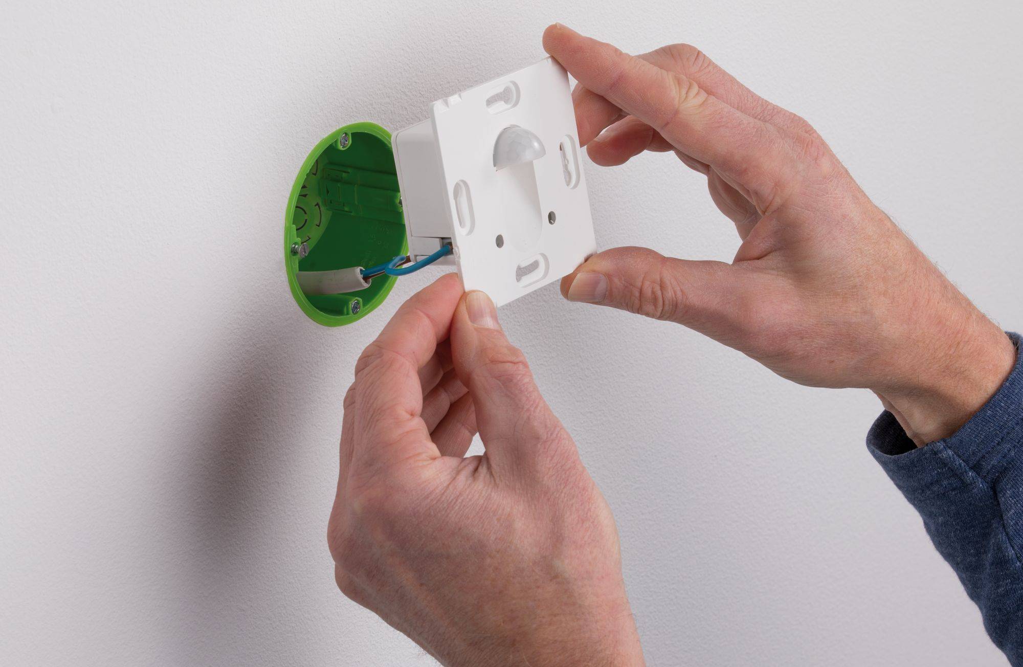 Eine Person installiert einen Bewegungssensor in einer Wanddose. Die Hände halten das Sensormodul an der Elektrikbox.