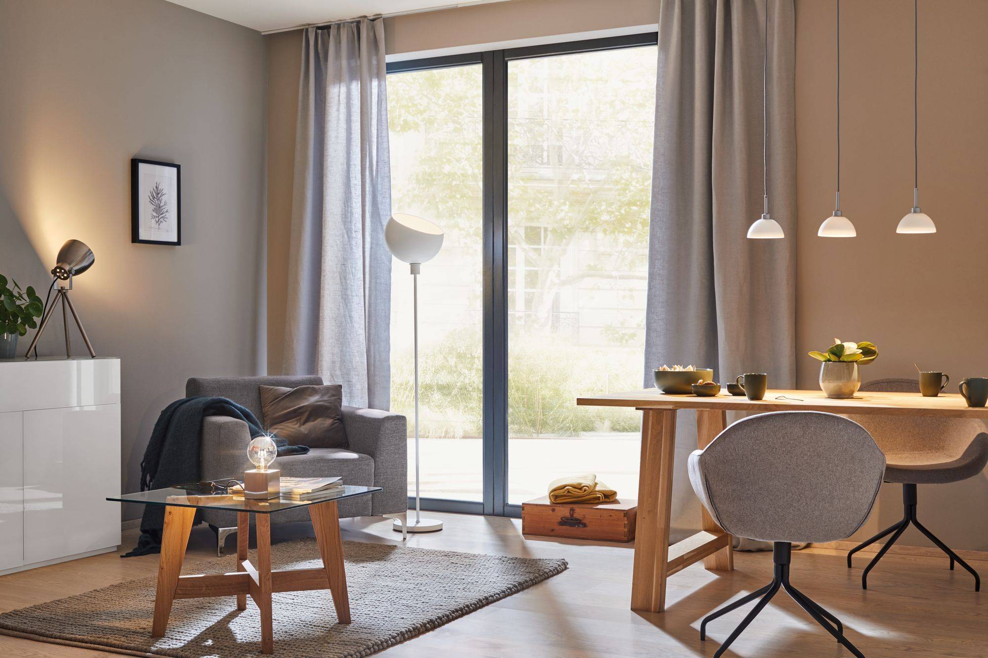 Moderner Wohnraum mit großem Fenster, Ledersessel, Tischlampe, Holztisch mit Stühlen und Hängelampen; helle und einladende Atmosphäre.