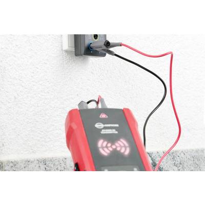 Ein Spannungsprüfer ist in eine Steckdose eingesteckt. Ein rotes Gerät mit Kabeln zeigt, dass der Stromkreis aktiv ist.