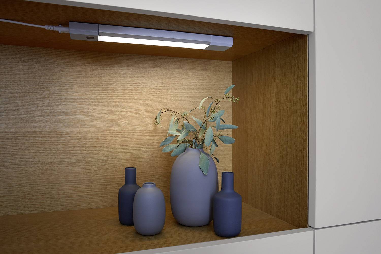 Ein beleuchtetes Holzregal mit vier Vasen in verschiedenen Formen und Größen. Eine Vase enthält getrocknete Zweige mit Blättern.