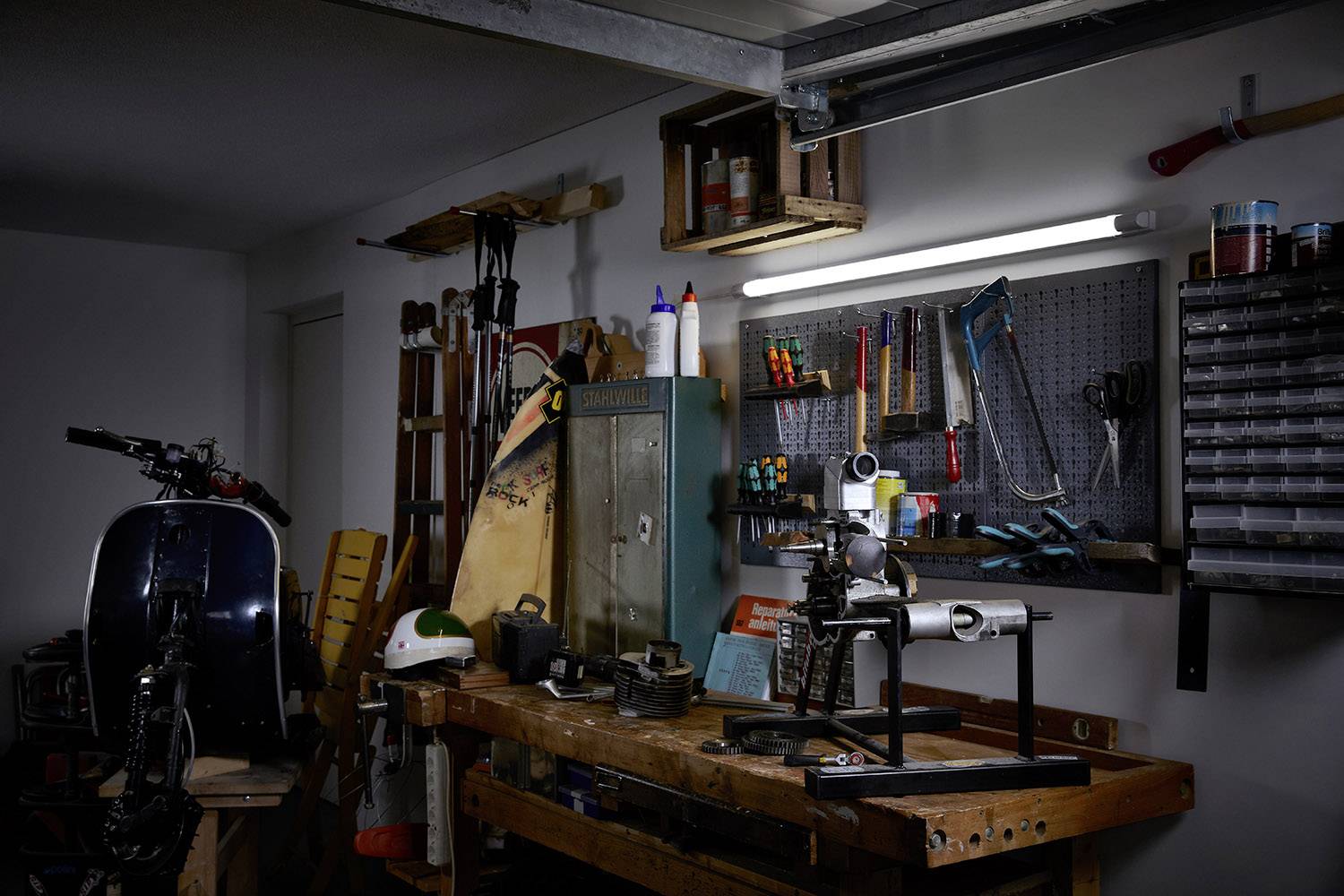 Eine Werkstatt mit Werkbank, Werkzeug an der Wand, einem Fahrradrahmen in einer Halterung, und einem Schrank mit Farbdosen.